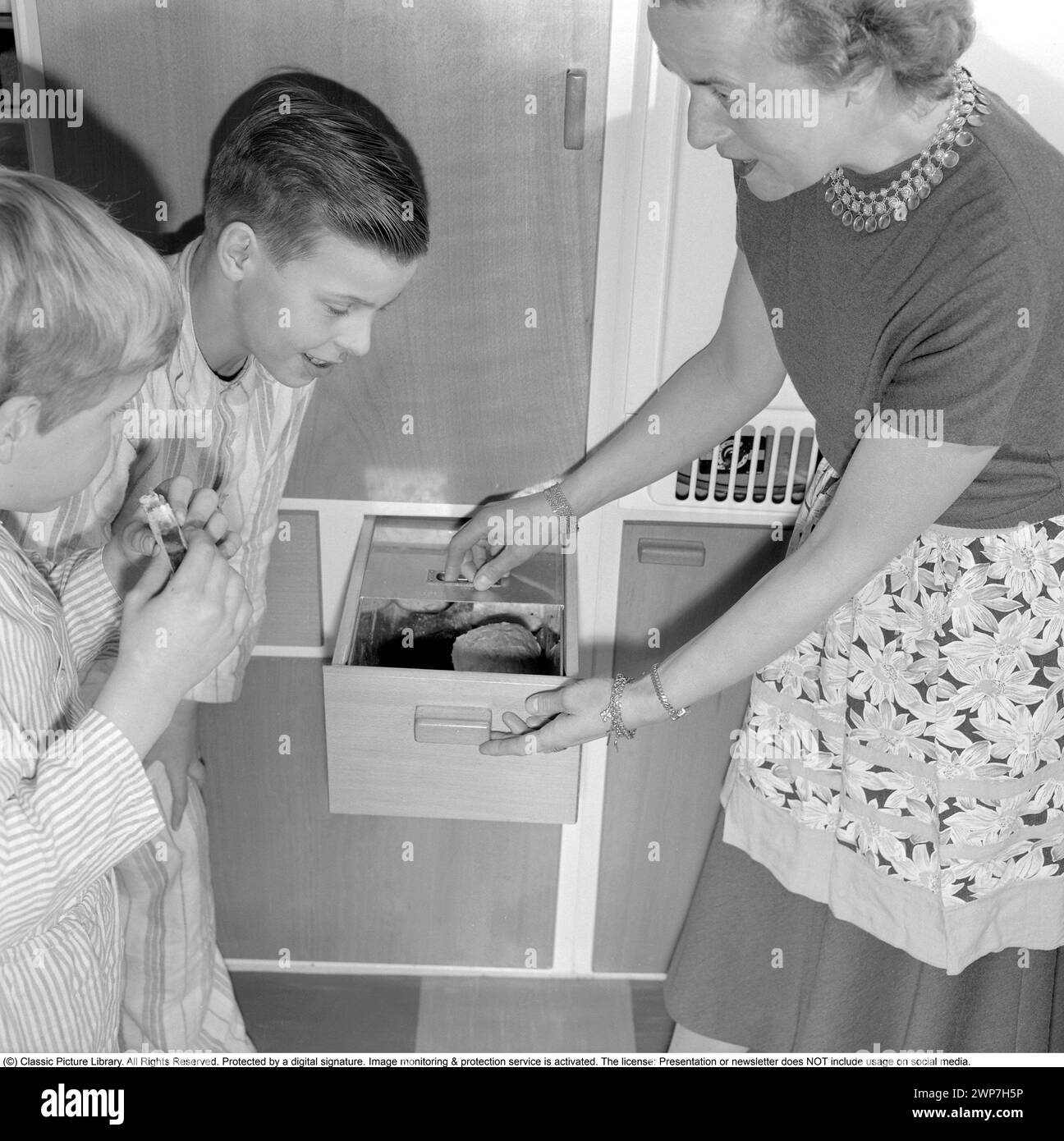 Desayuno en los años 1950 Dos chicos en pijama preparándose en la cocina. Uno de ellos comiendo un sándwich. Su madre les muestra el práctico cajón donde se guarda el pan. 1956. Conard ref 3167 Foto de stock