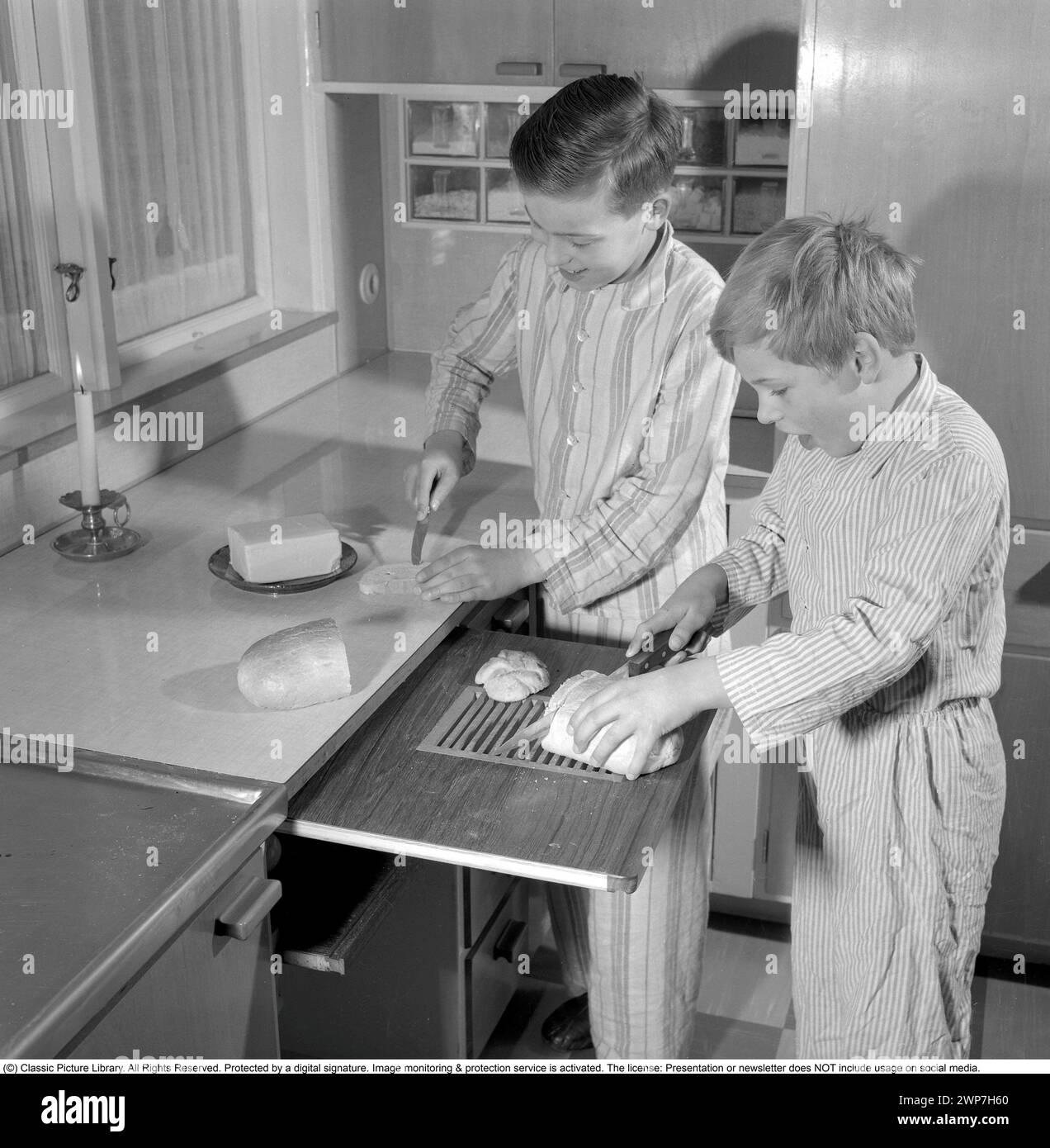 Desayuno en los años 1950 Dos niños están de pie en pijama preparando el desayuno. Uno de ellos corta el pan en la tabla de cortar y uno extiende la mantequilla en el sándwich. 1956. Conard ref 3167 Foto de stock