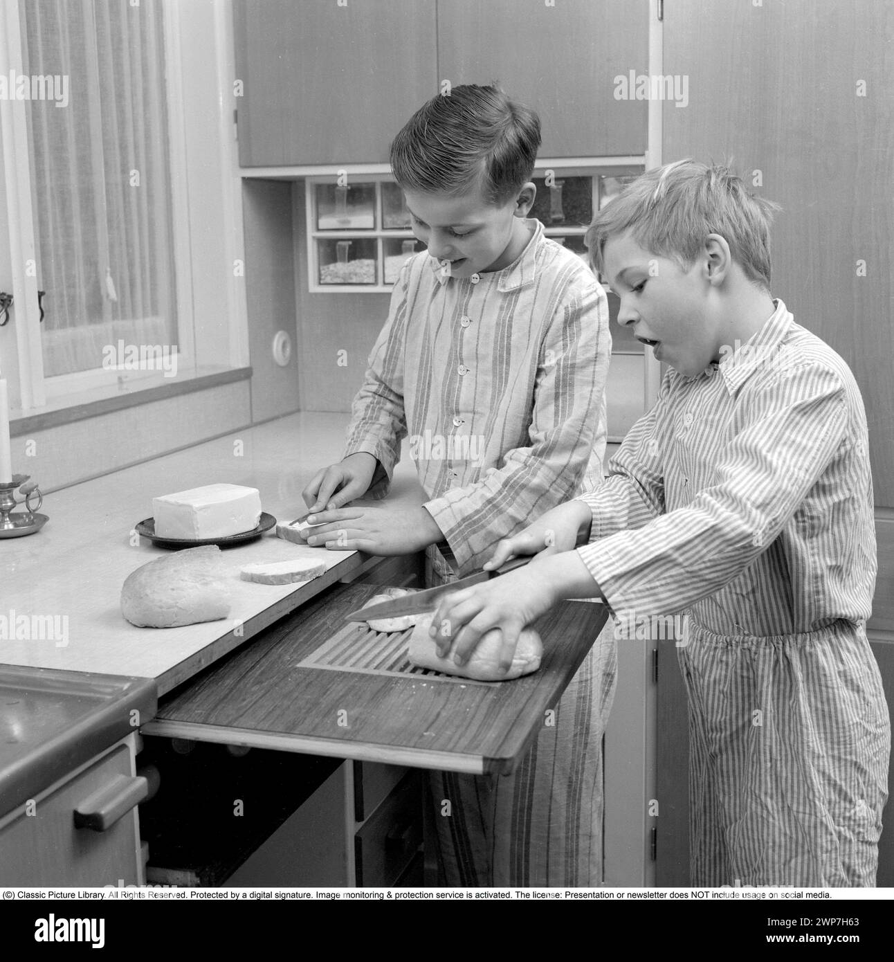 Desayuno en los años 1950 Dos niños están de pie en pijama preparando el desayuno. Uno de ellos corta el pan en la tabla de cortar y uno extiende la mantequilla en el sándwich. 1956. Conard ref 3167 Foto de stock