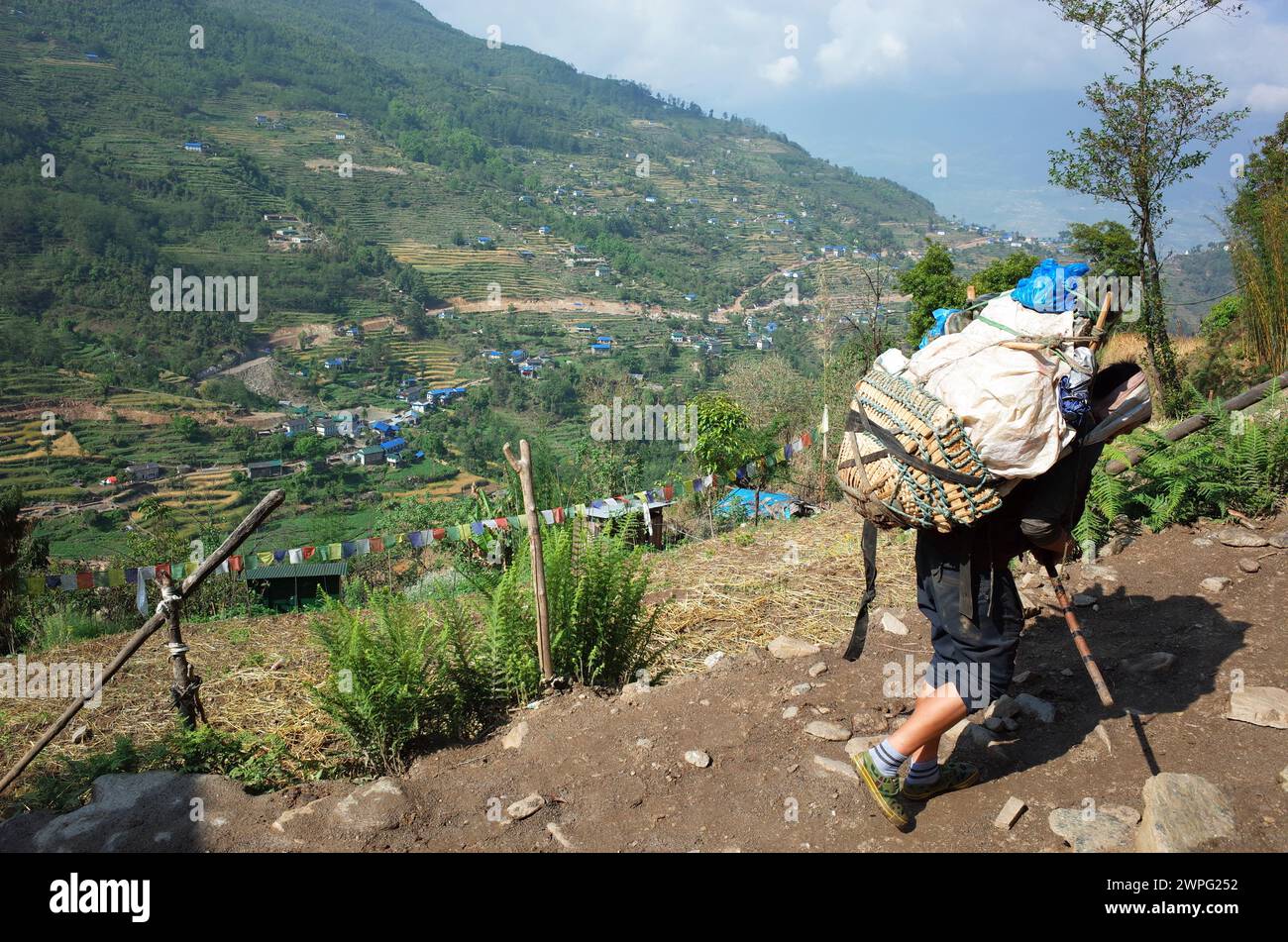 Porteador que lleva carga pesada en la cesta de las montañas verdes del Himalaya con vista sobre el pueblo de Karikhola, Solukhumbu, región del Everest, Nepal Foto de stock