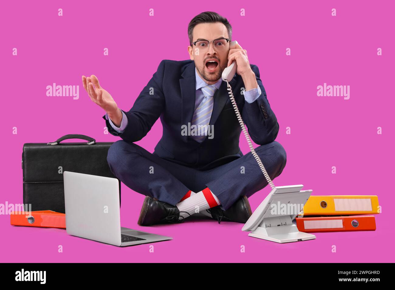 Hombre de negocios divertido con el ordenador portátil, las carpetas y el maletín hablando por teléfono sobre fondo púrpura Foto de stock