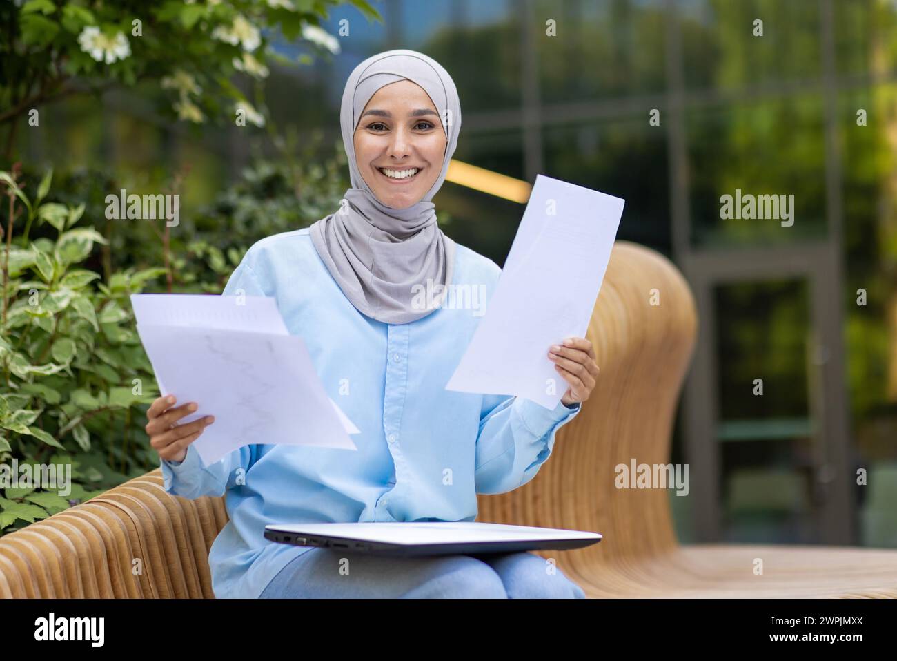 Una mujer musulmana profesional con un pañuelo sosteniendo papeles, mostrando confianza y una cálida sonrisa, sentada en un banco al aire libre. Foto de stock