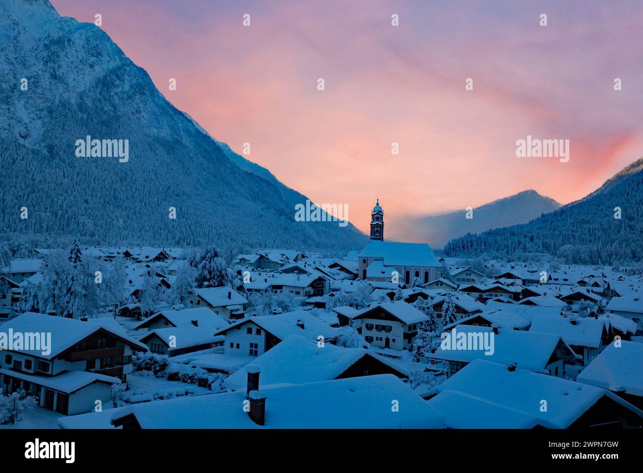 La mañana de invierno en Mittenwald antes del amanecer, la llamativa torre de la iglesia, el amanecer, los techos de las casas cubiertas de nieve y las montañas Foto de stock