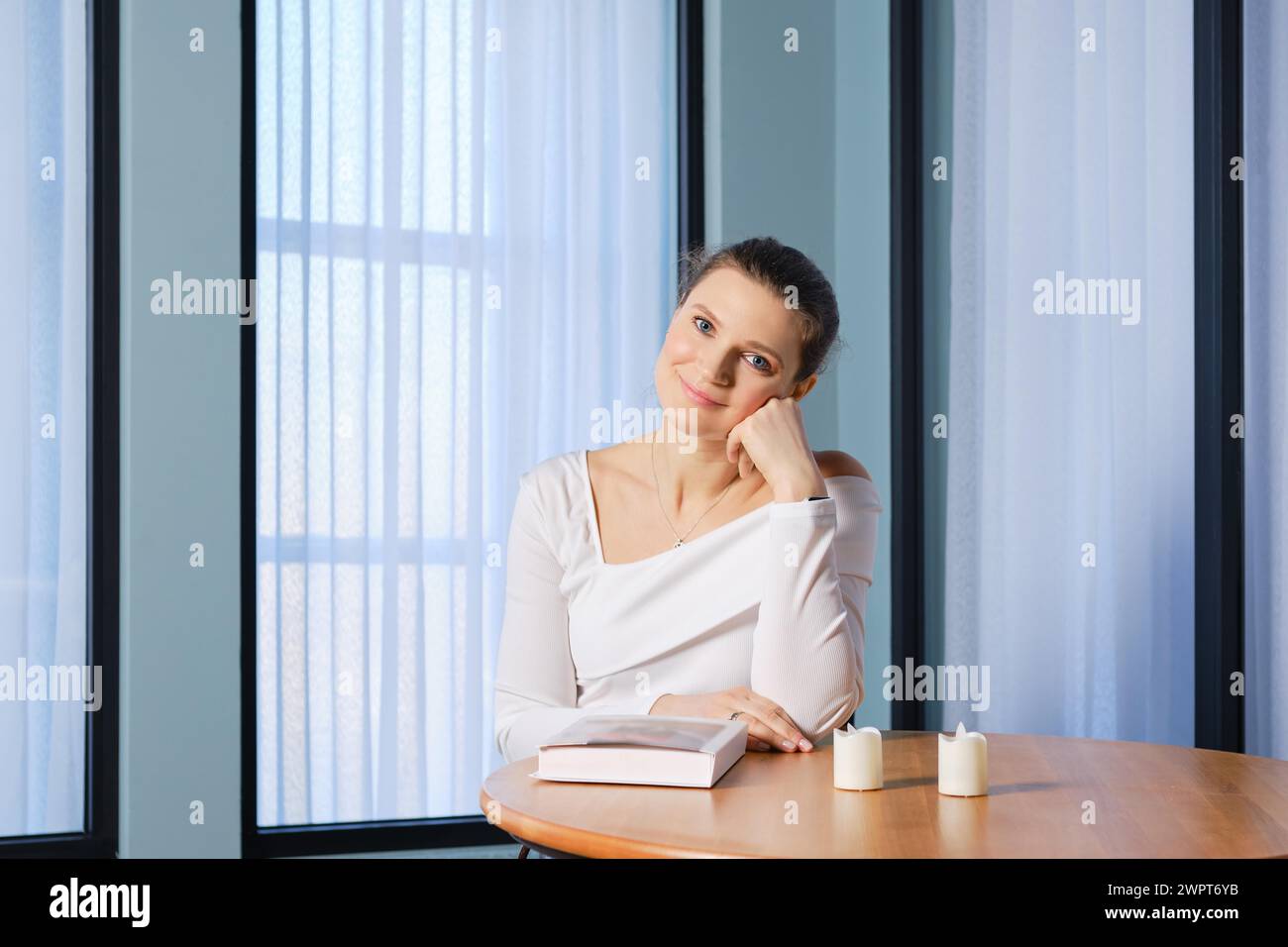 Mujer soñadora se sienta en una mesa de madera con un libro, sostiene la cabeza con el puño y se ve con una sonrisa amable Foto de stock