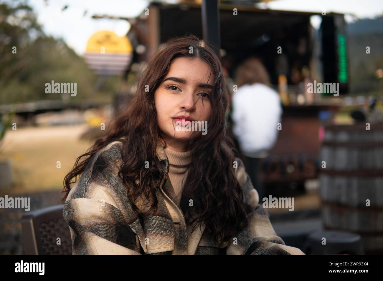 Una joven latina con una sonrisa cálida se sienta cómodamente en un ambiente de cafetería al aire libre, exudando elegancia casual Foto de stock