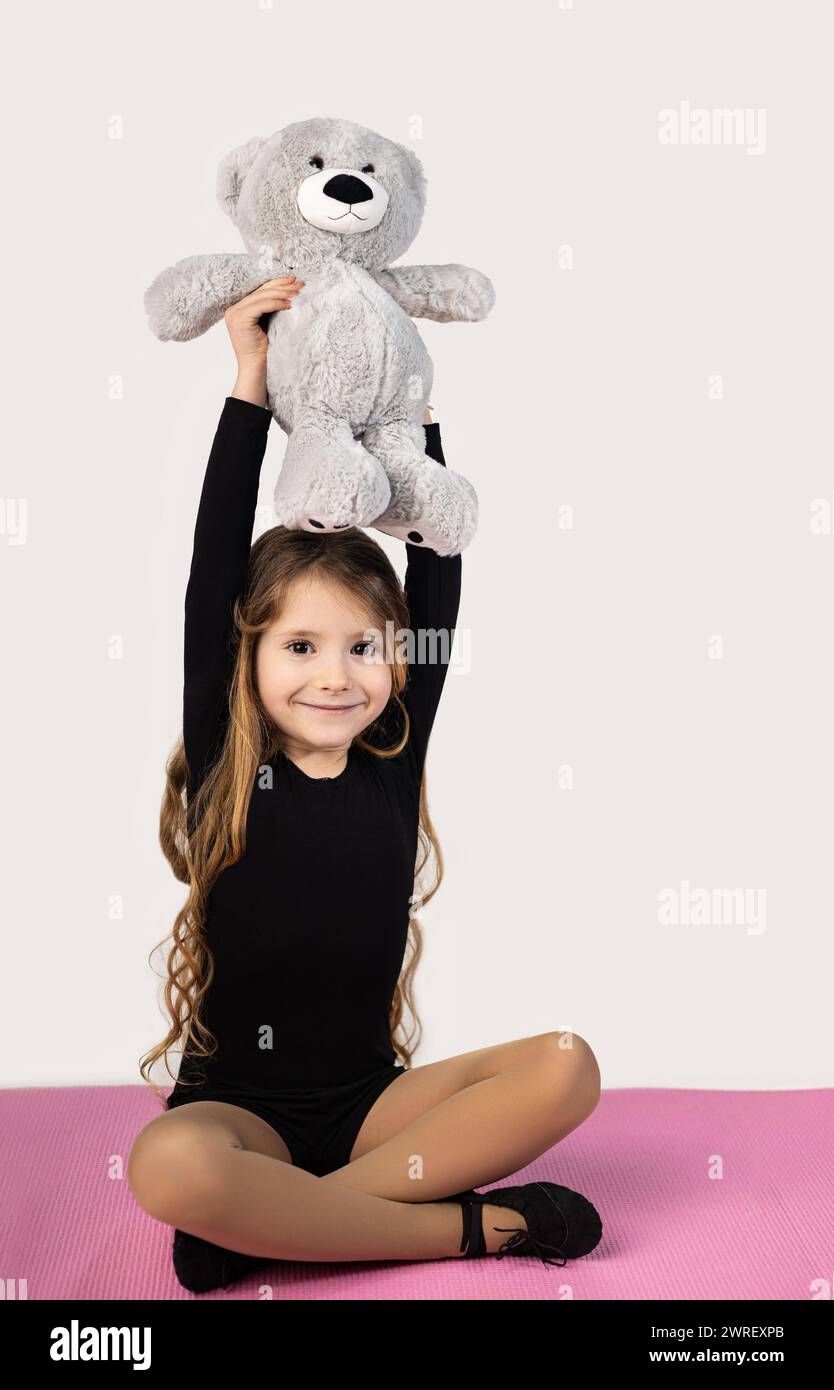 Foto vertical de una niña gimnasta en una esterilla de yoga con un traje negro, sosteniendo sus manos en alto con un oso de peluche. Foto de alta calidad Foto de stock