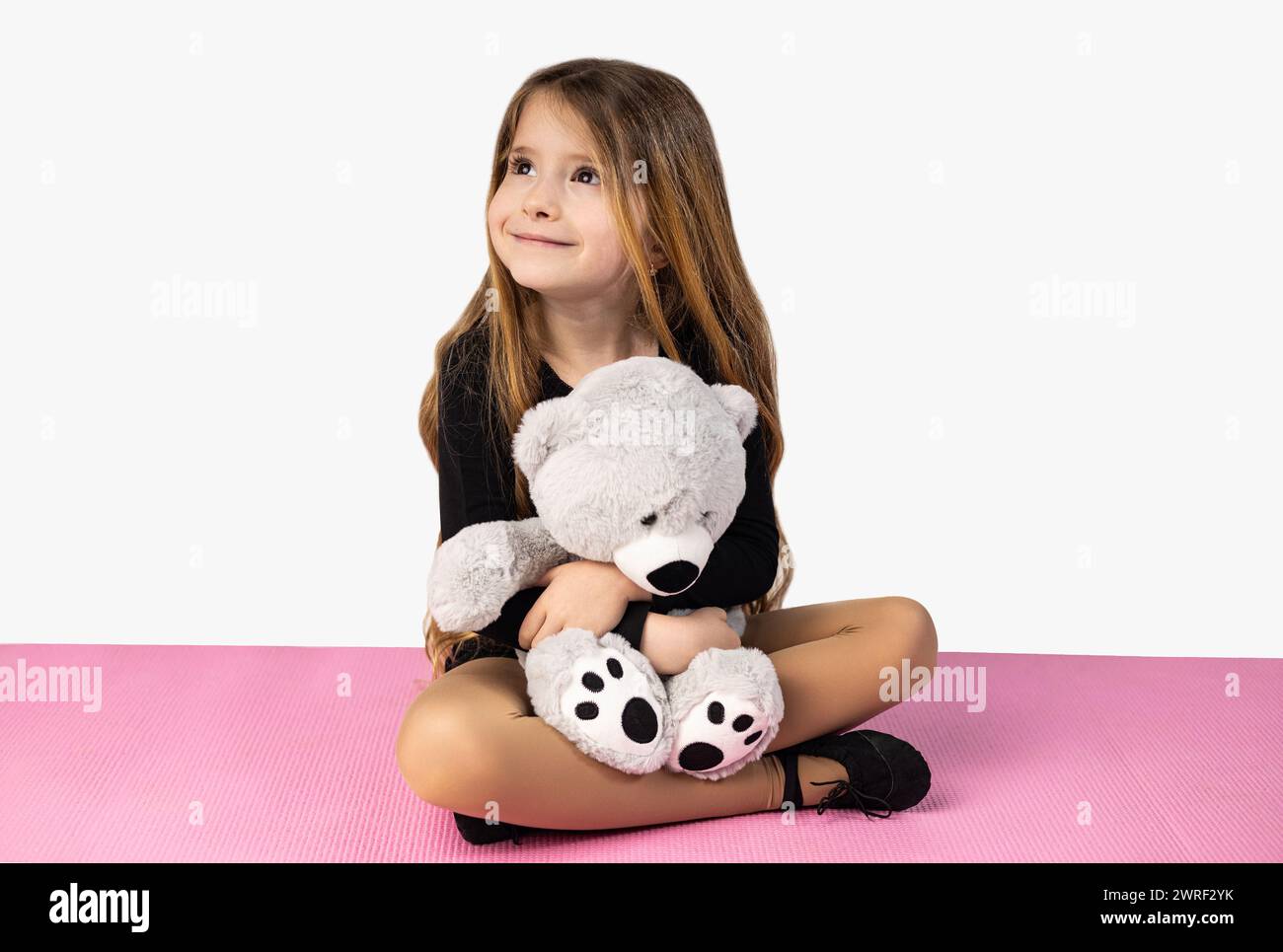 Niña sonriente vestida con ropa negra de gimnasio, sosteniendo un oso de peluche gris en sus brazos, sentada en una esterilla de yoga rosa, aislada en un backgr de estudio blanco Foto de stock