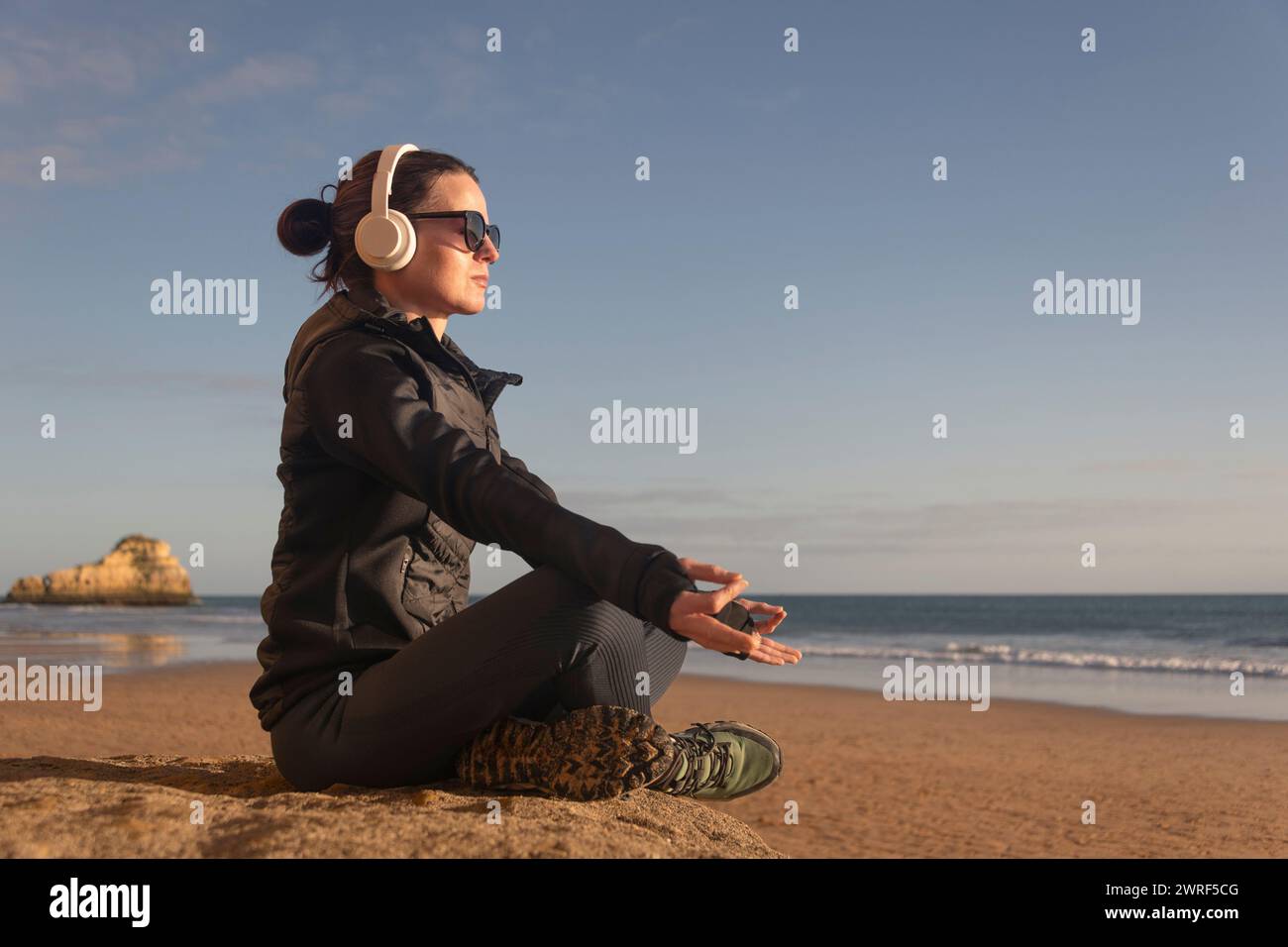 Mujer deportiva sentada meditando junto al mar, usando auriculares y gafas de sol. Foto de stock