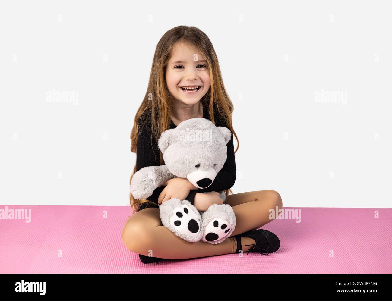 Niña sonriente vestida con ropa negra de gimnasio, sosteniendo un oso de peluche gris en sus brazos, sentada en una esterilla de yoga rosa, aislada en un backgr de estudio blanco Foto de stock