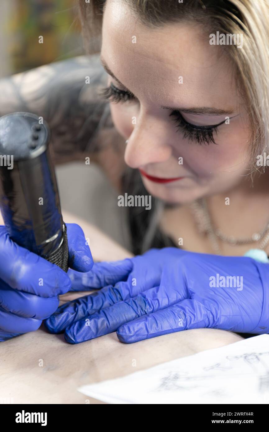 Foto vertical Un primer plano íntimo de una artista del tatuaje mientras se enfoca intensamente en entintar un diseño, sus guantes azules resaltando el tono del tatuaje. Con Foto de stock