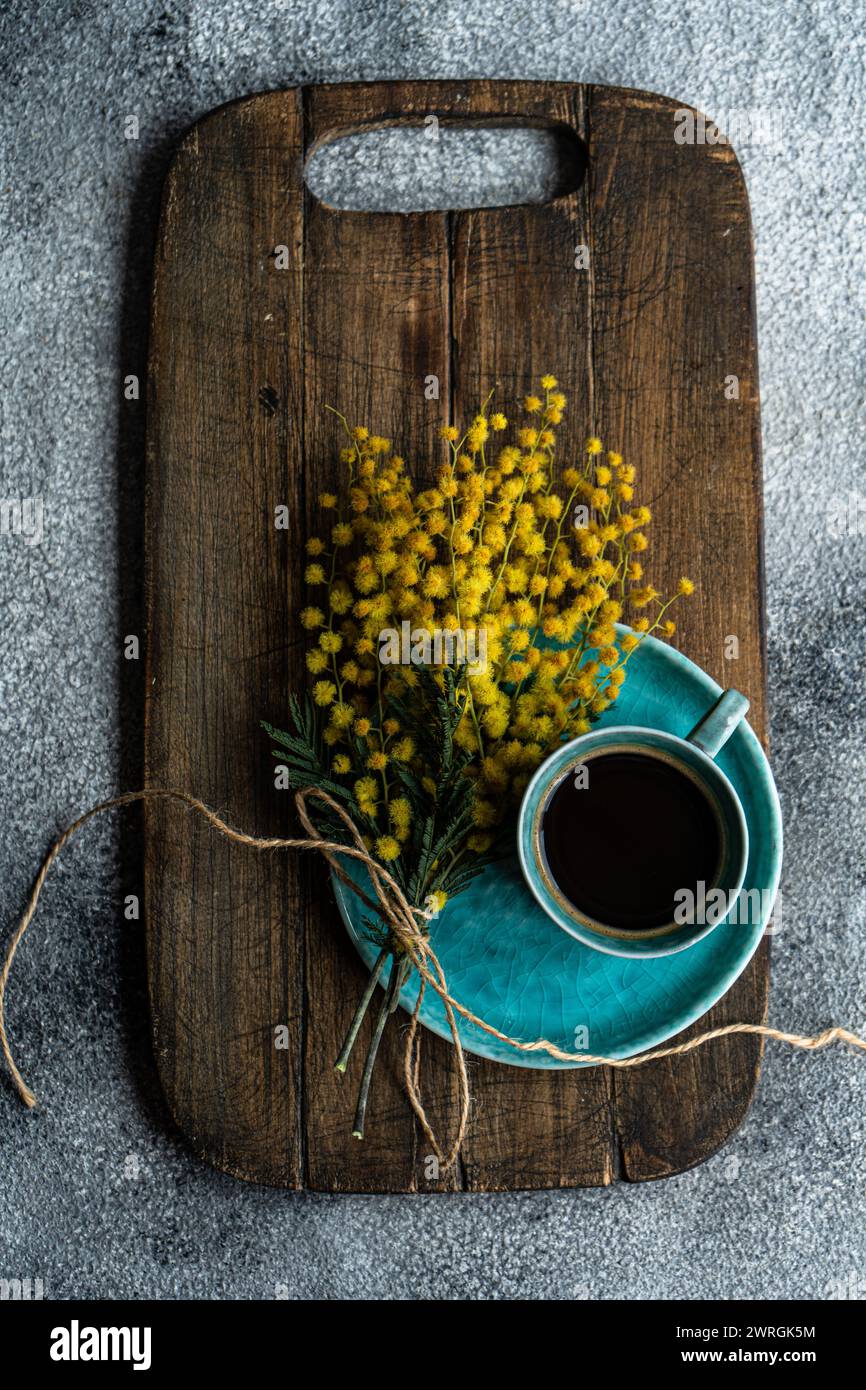 Vista de arriba de una taza de café negro y un montón de flores de mimosa amarilla en una tabla de cortar de madera Foto de stock
