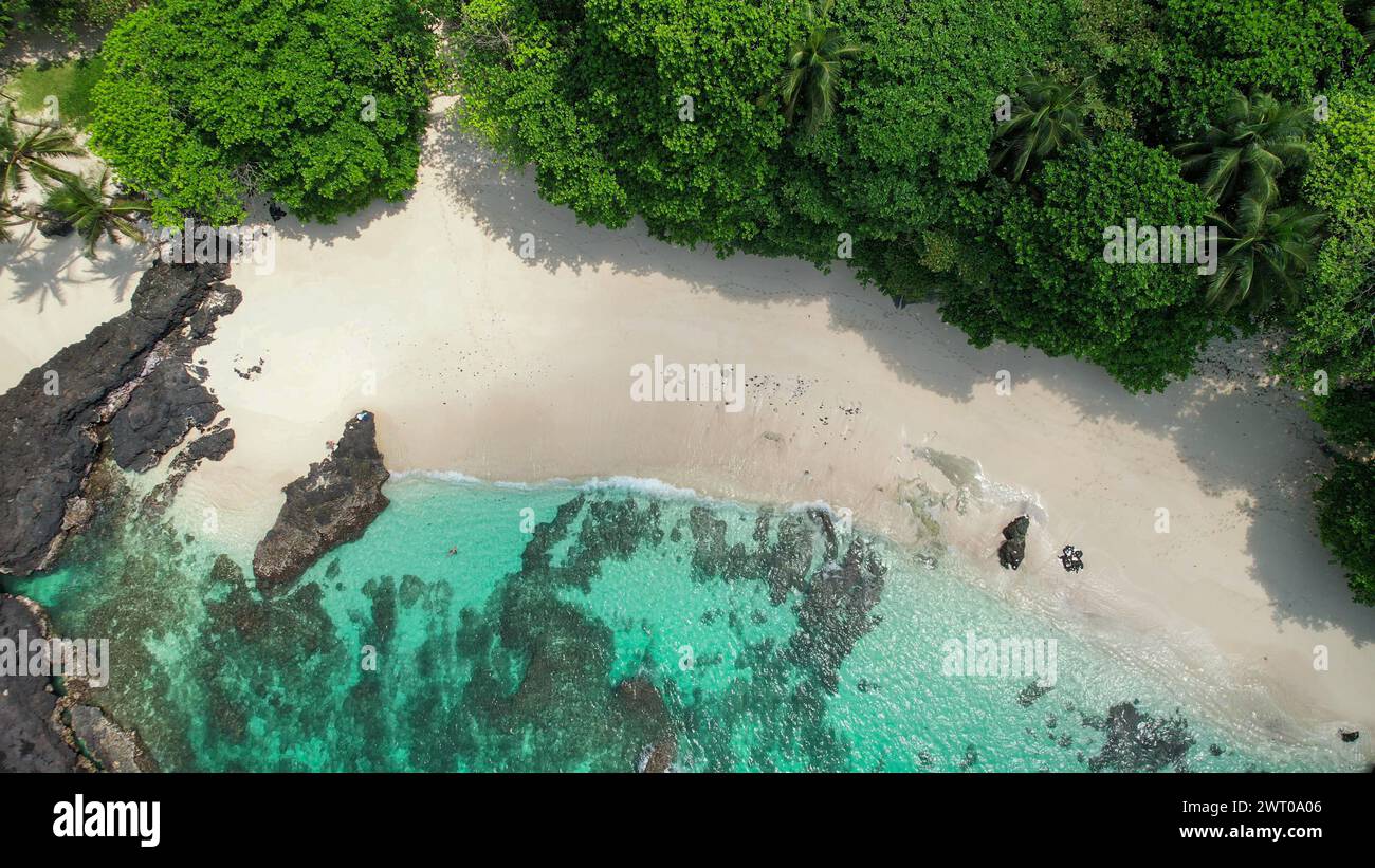 Una playa de arena blanca rodeada de exuberantes árboles verdes, mar turquesa y cielos claros, Ilheu das Rolas islote en Santo Tomé, África Foto de stock