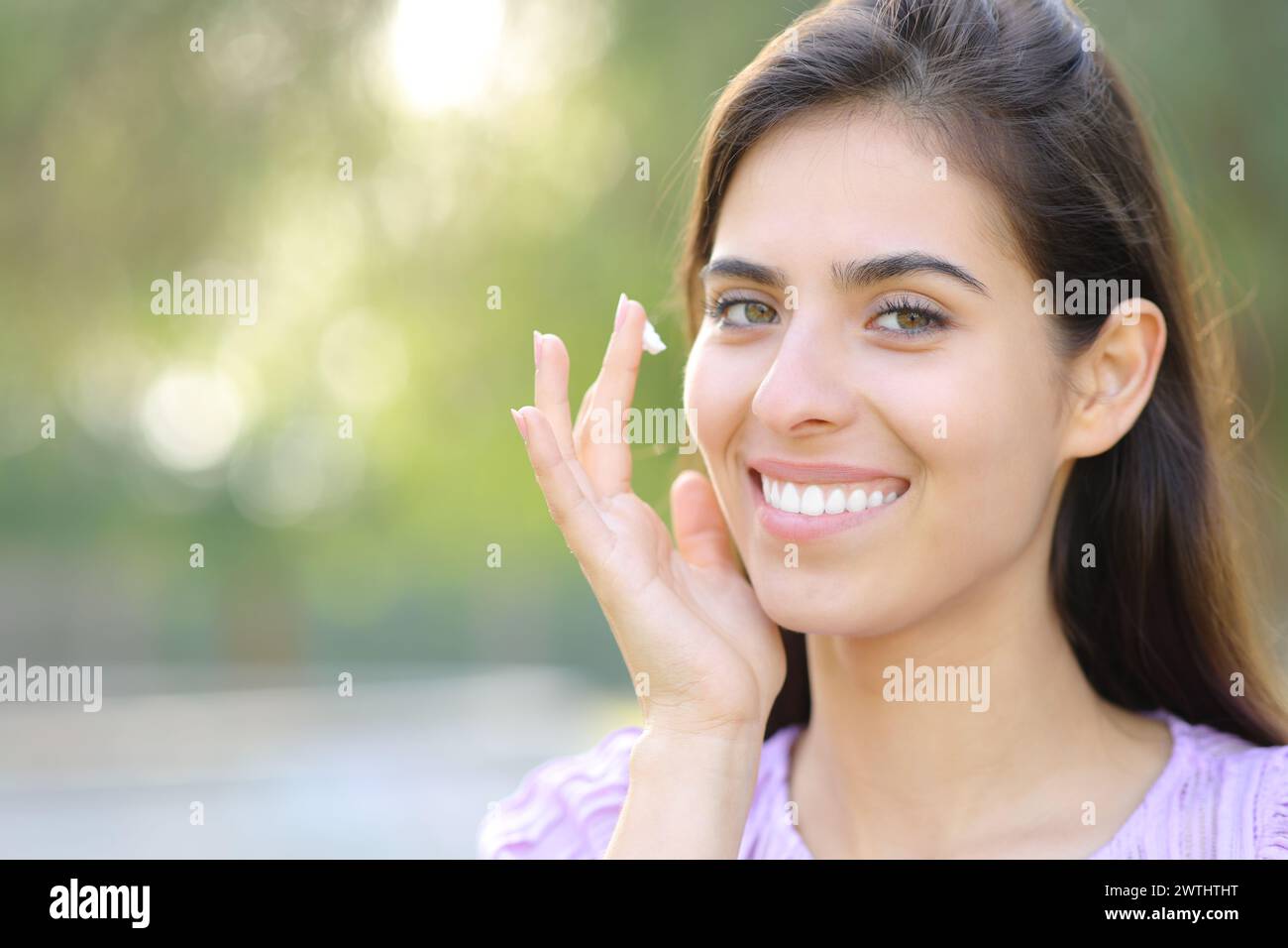 Retrato de una mujer feliz aplicando humectante mirándote en un parque Foto de stock