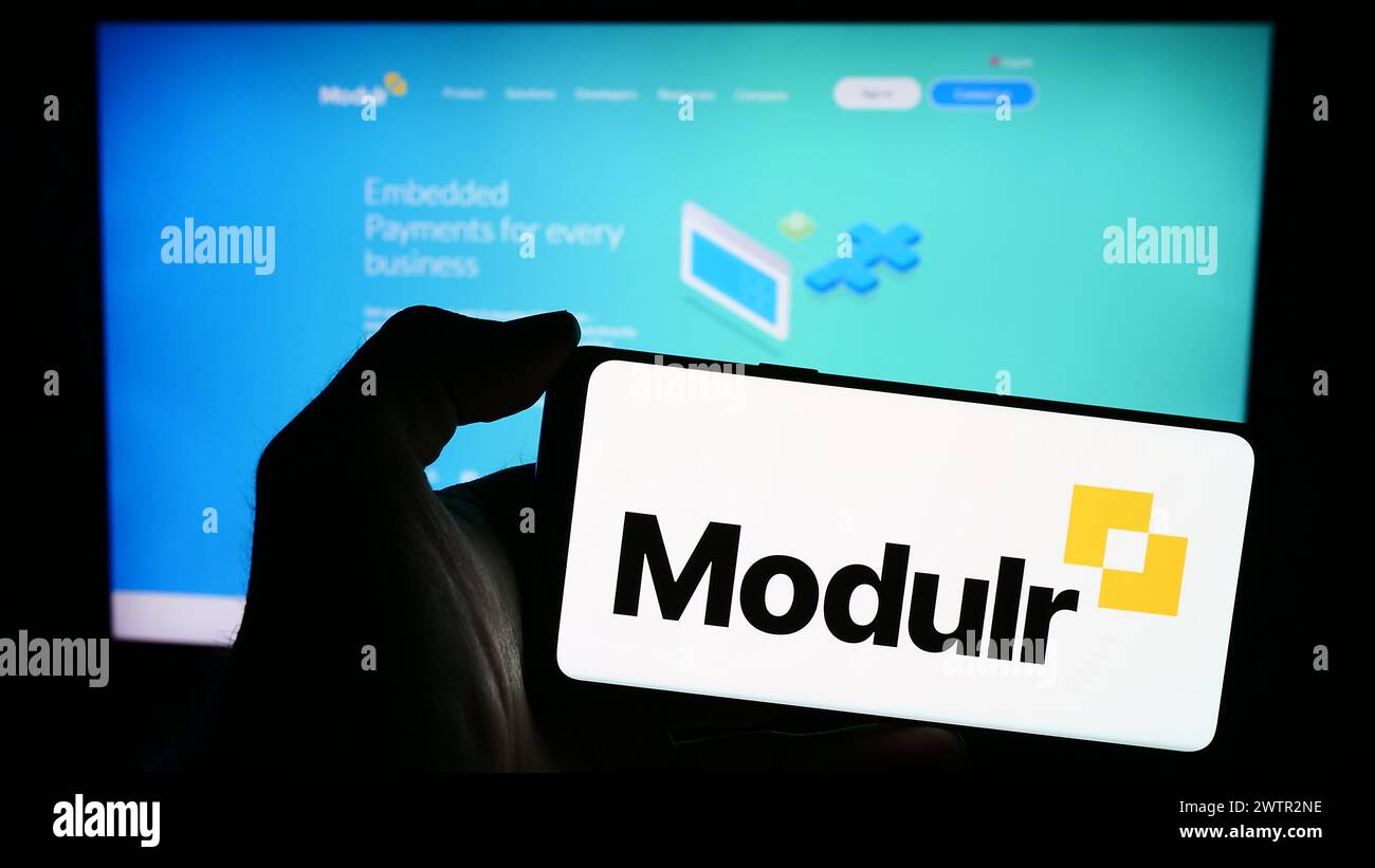 Persona que sostiene el teléfono móvil con el logotipo de la compañía de pagos Modulr Finance BV frente a la página web del negocio. Enfoque en la pantalla del teléfono. Foto de stock