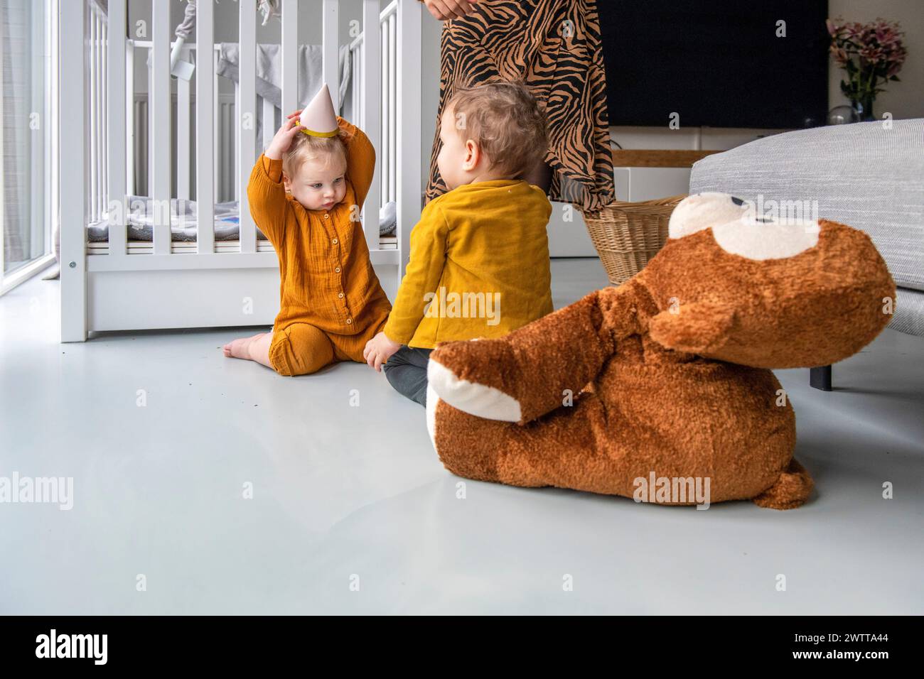 Dos niños pequeños jugando en el suelo con un gran animal de peluche en una habitación iluminada. Foto de stock