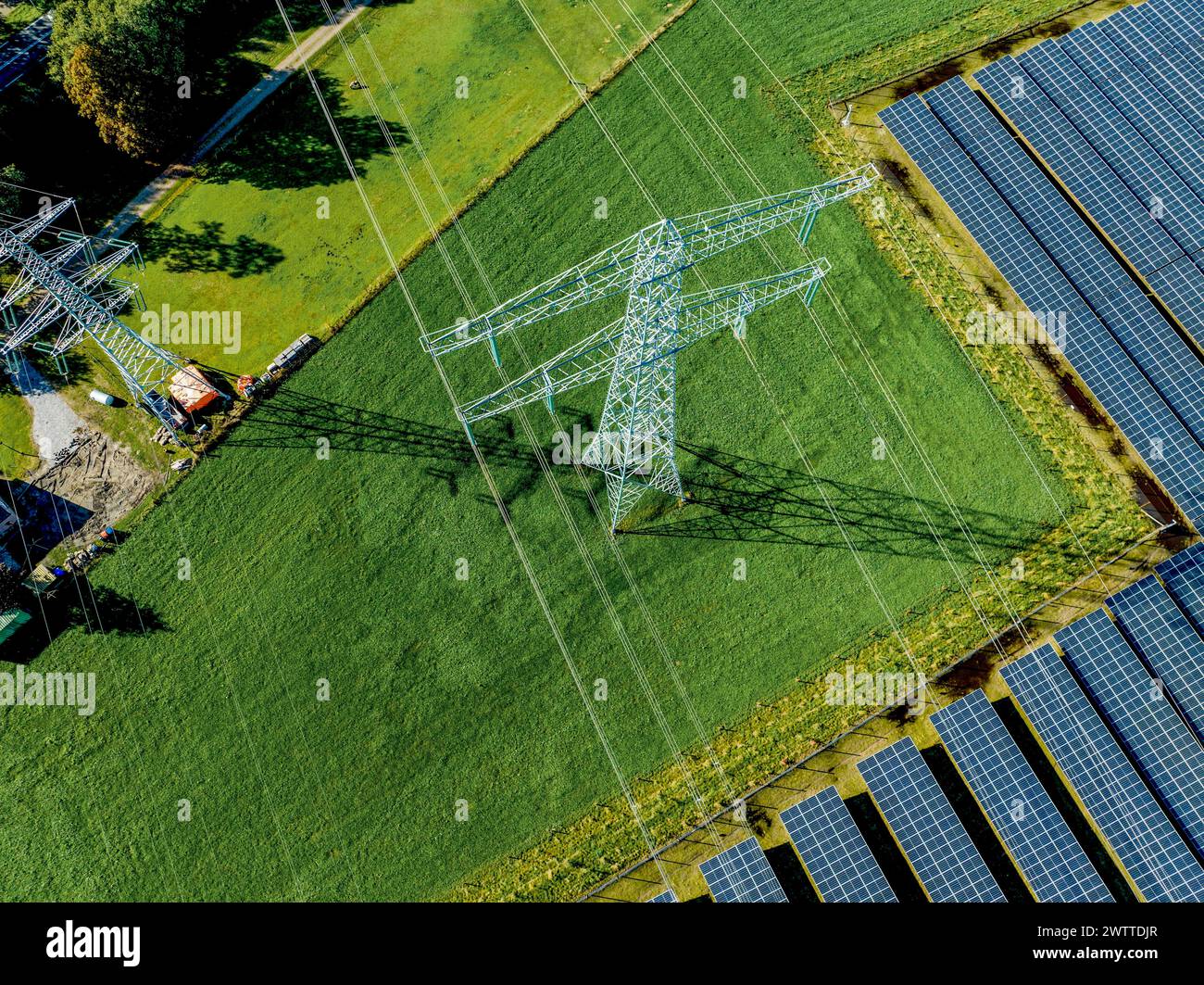 Vista aérea de paneles solares junto a un pilón de electricidad en exuberantes campos verdes Foto de stock