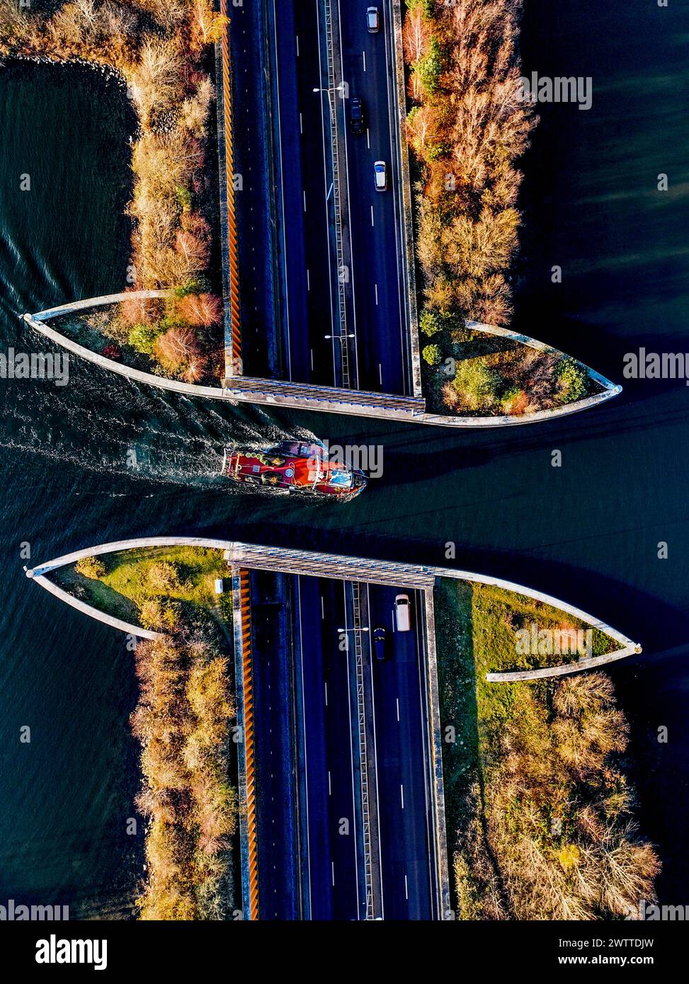 Vista aérea de un barco navegando bajo un puente sobre un río iluminado por el sol, flanqueado por caminos y árboles de otoño. Foto de stock