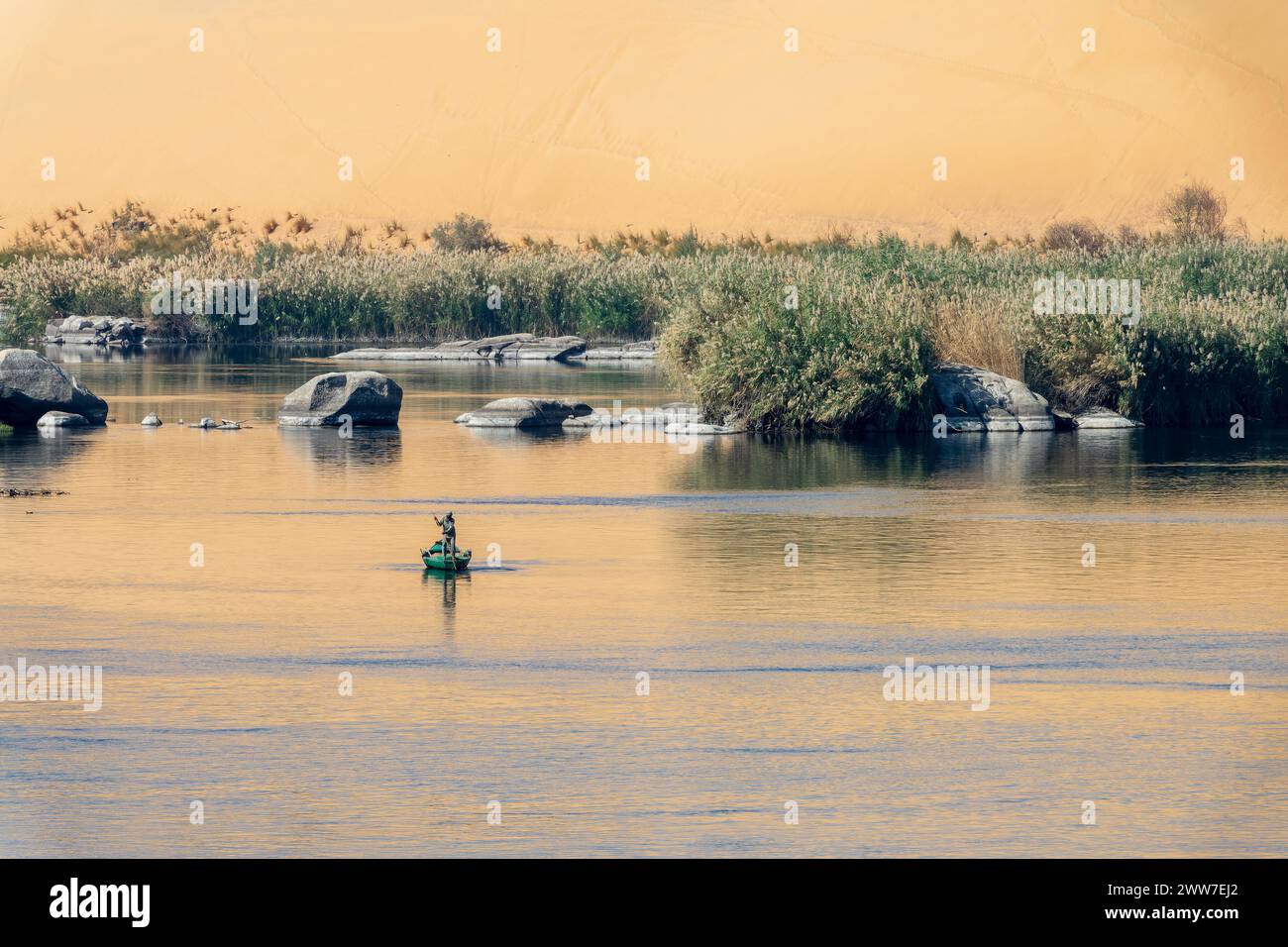 Pescador en un bote de remos en el río Nilo al sol, paisaje escénico con rocas en el agua y reflejos de dunas de arena en Asuán, Egipto Foto de stock