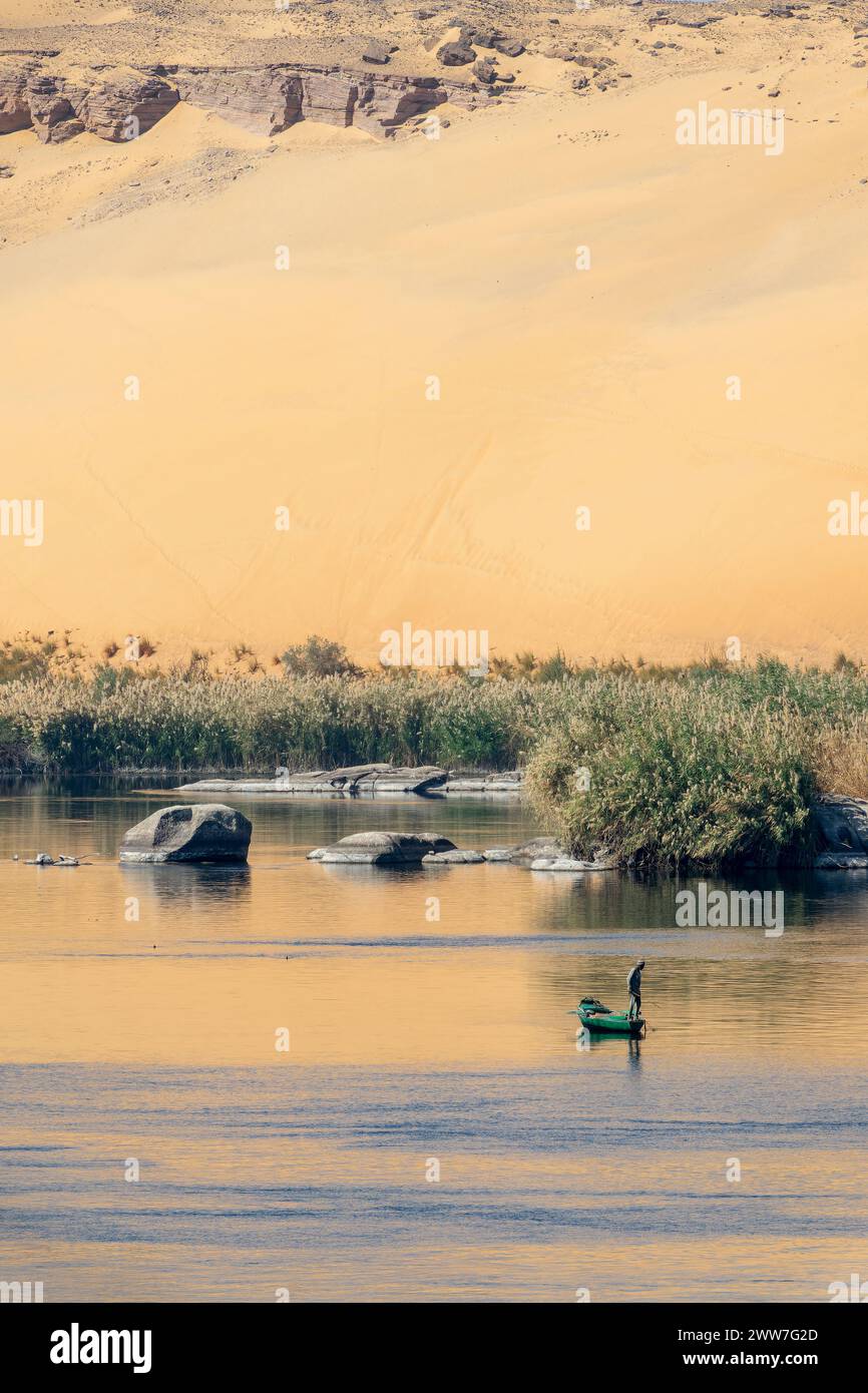 Pescador en un bote de remos en el río Nilo al sol, paisaje escénico con rocas en el agua y reflejos de dunas de arena en Asuán, Egipto Foto de stock