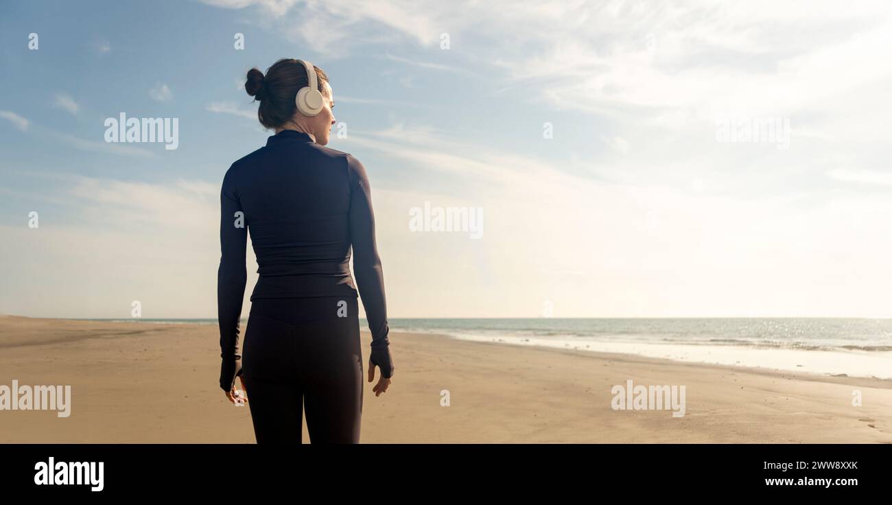 vista trasera de una mujer deportiva caminando en una playa, pasos diarios, usando auriculares. Foto de stock