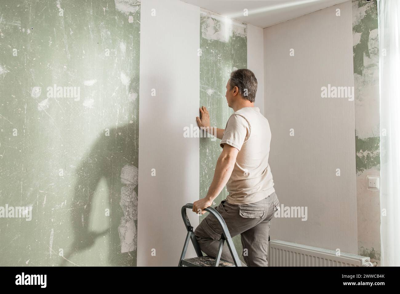 Hombre en escalera de paso pegando papel pintado blanco en la sala de estar en casa Foto de stock