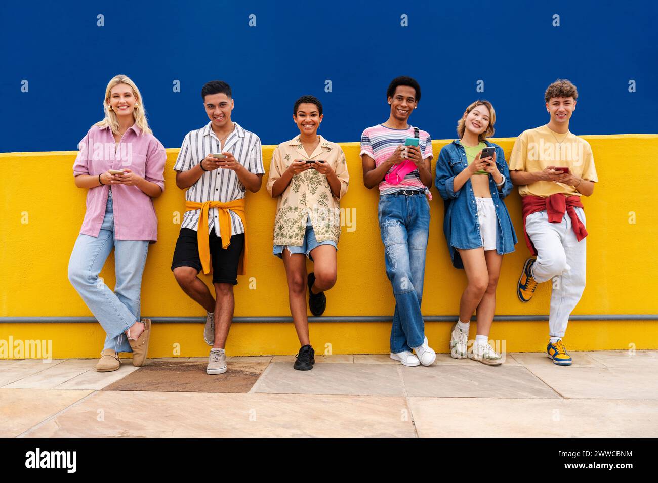 Grupo de amigos apoyados contra la pared amarilla usando sus teléfonos inteligentes Foto de stock