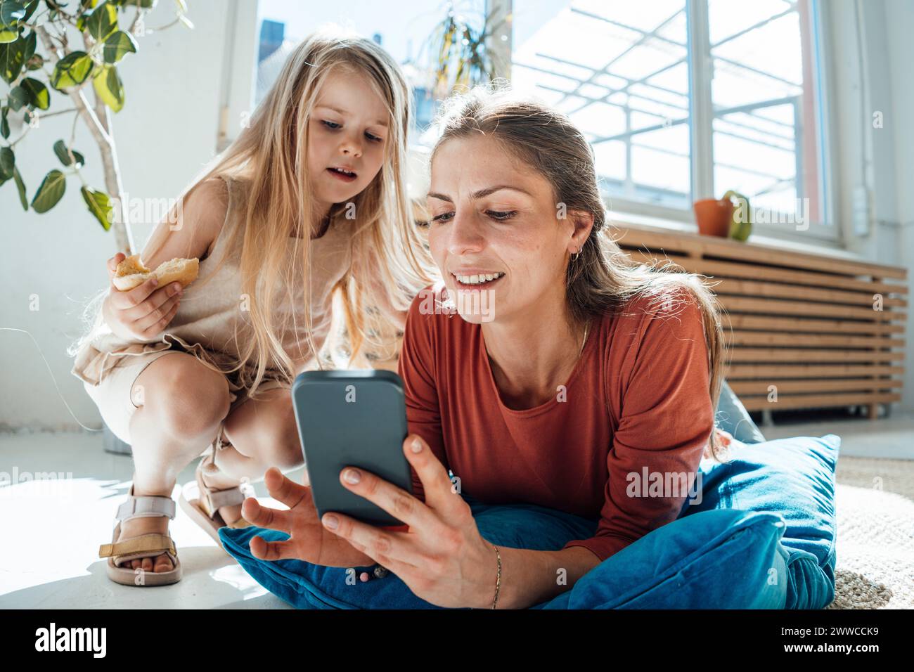 Mujer sonriente usando el teléfono inteligente con la hija que sostiene el bocadillo en casa Foto de stock