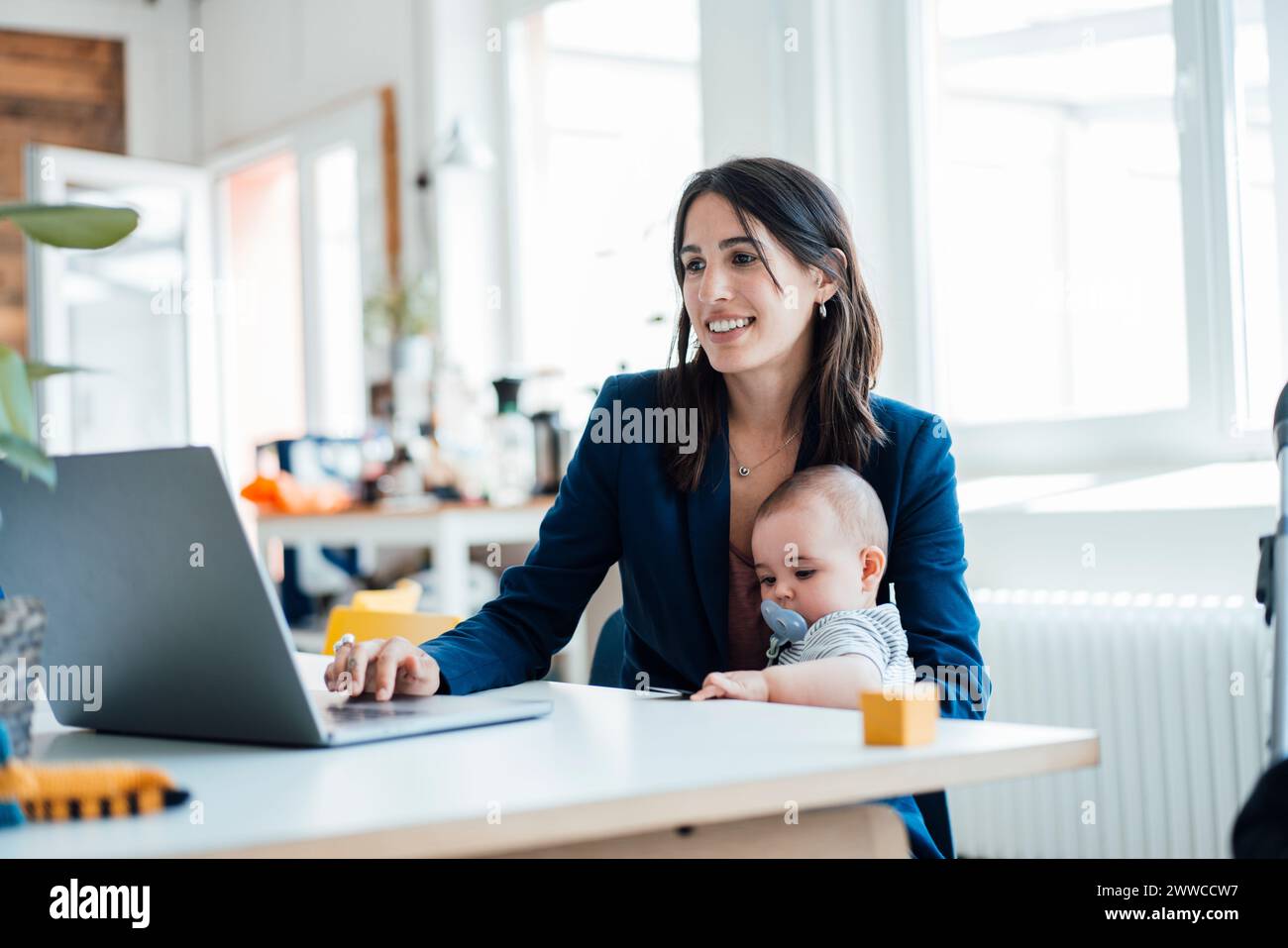 Empresaria sonriente que se sienta con la muchacha del bebé usando el ordenador portátil en casa Foto de stock