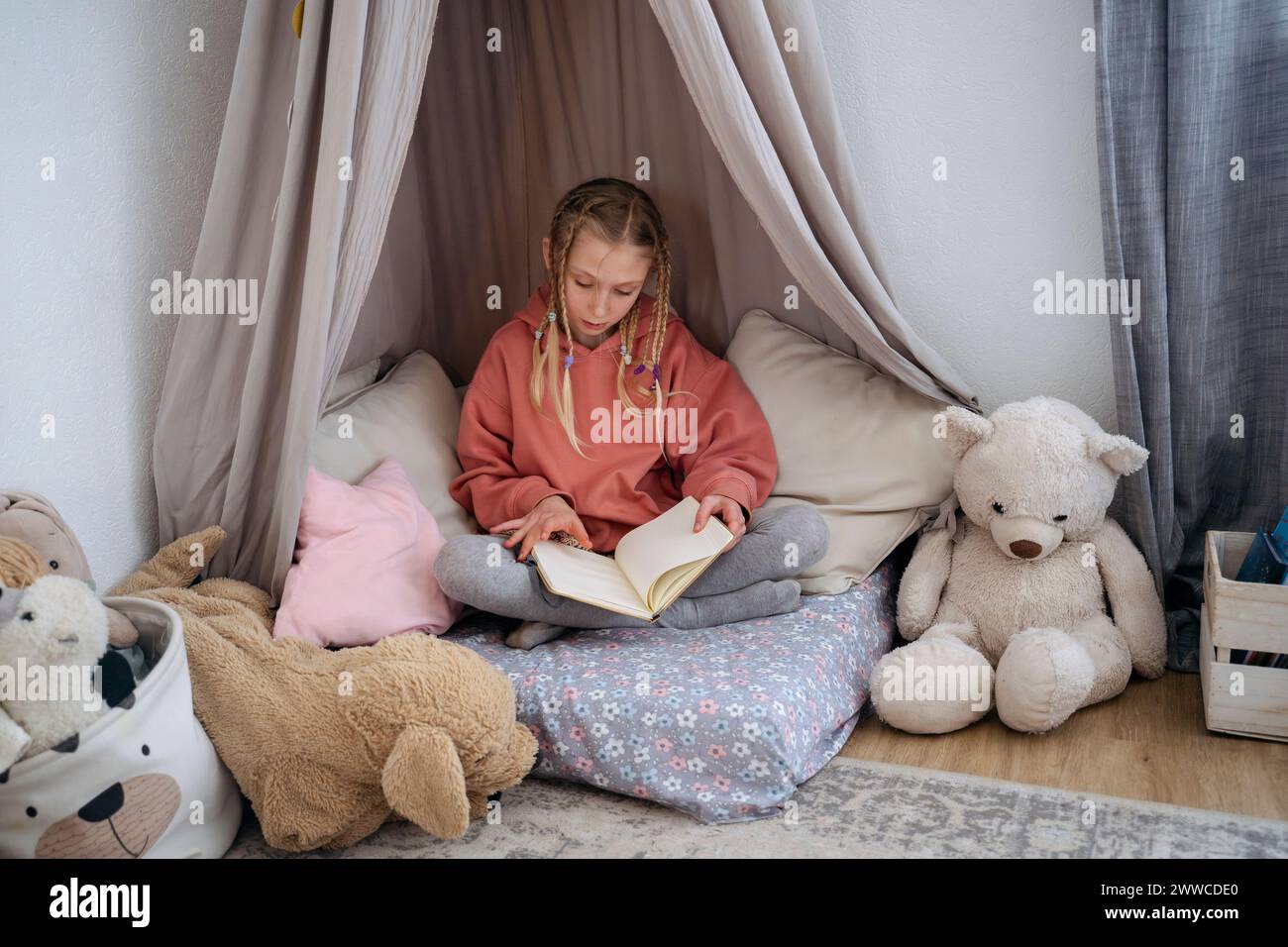 Muchacha con el libro de lectura de las coletas en canopy gris en casa Foto de stock