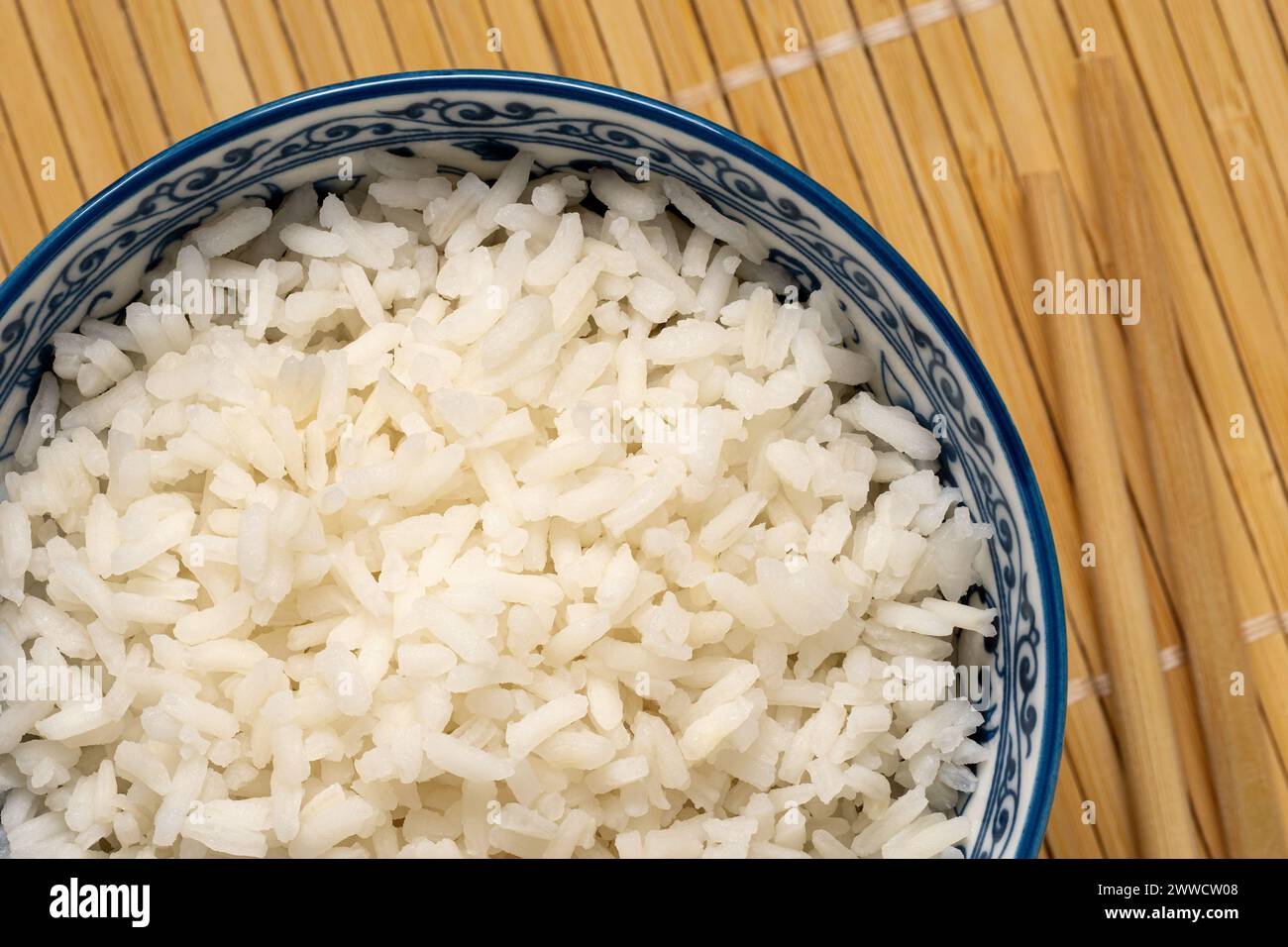 Detalle de arroz blanco cocido en un tazón asiático de cerámica blanca y azul junto a palillos aislados en mate de bambú ligero. Vista superior. Foto de stock