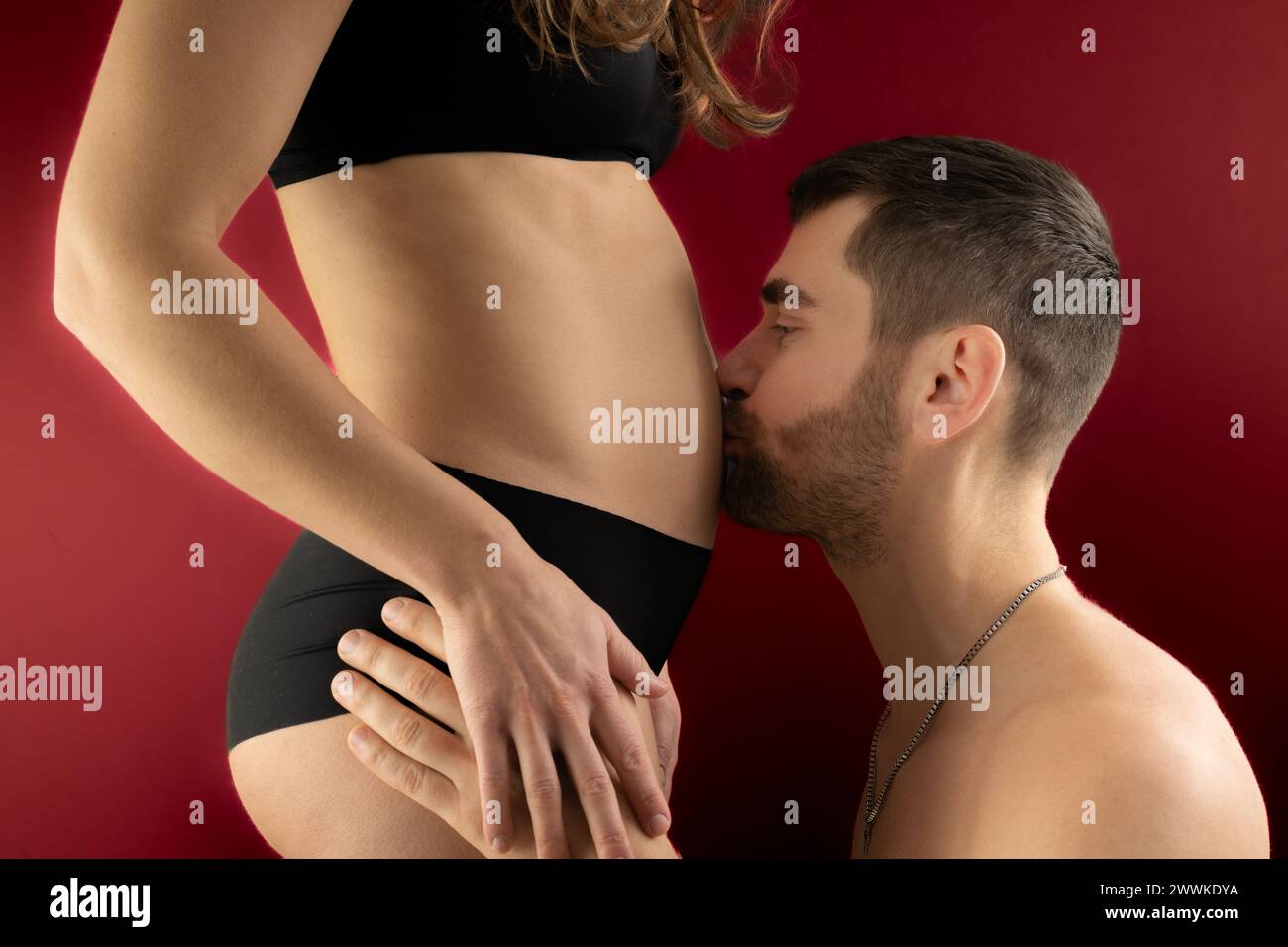 Descripción: Vista del vientre de la mujer embarazada con padre feliz besándolo. Embarazo Primer trimestre - semana 18. Vista lateral. Fondo rojo. Tiro brillante. Foto de stock