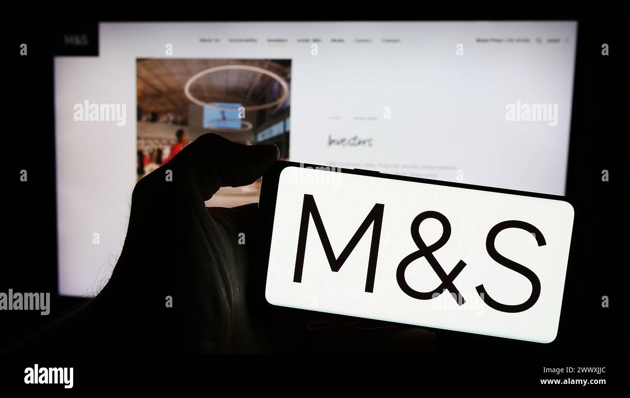 Persona que sostiene el teléfono móvil con el logotipo de la compañía minorista británica Marks y Spencer Group plc delante de la página web del negocio. Enfoque en la pantalla del teléfono. Foto de stock