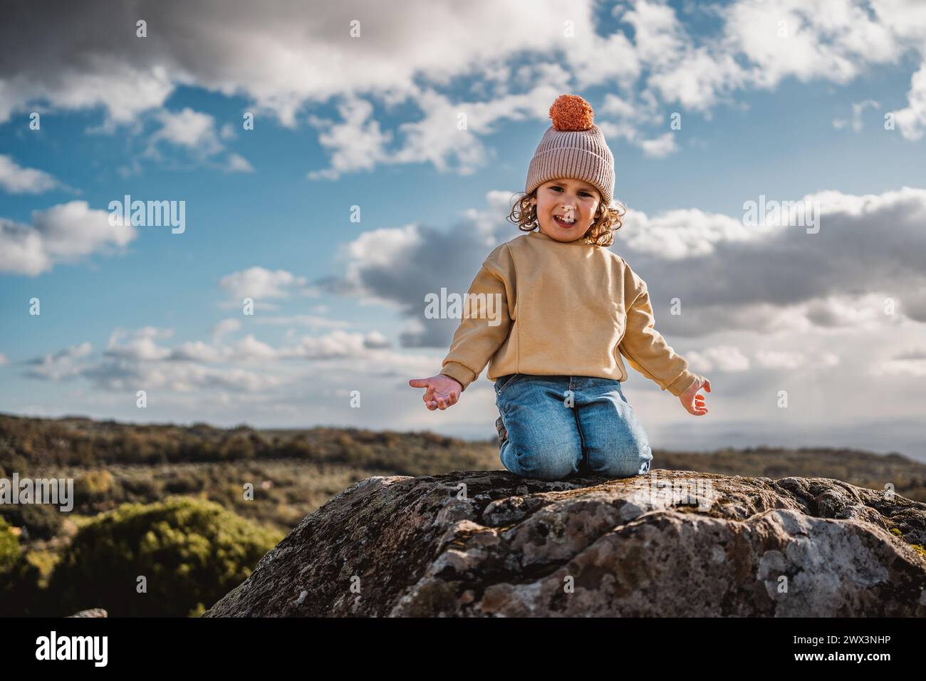 niño de moda con el sombrero jugando y feliz, preguntándose, en la cima de una roca, en un castillo de piedra con vista panorámica de las montañas, en el día soleado a Foto de stock