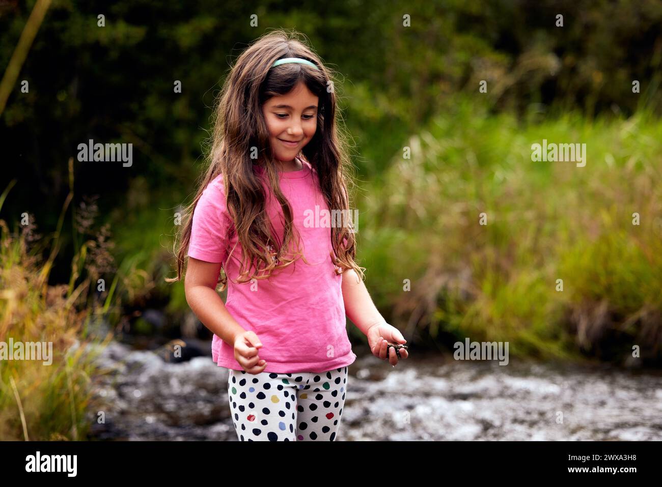 Chica sonriente sosteniendo guijarros mientras juega cerca de la corriente Foto de stock