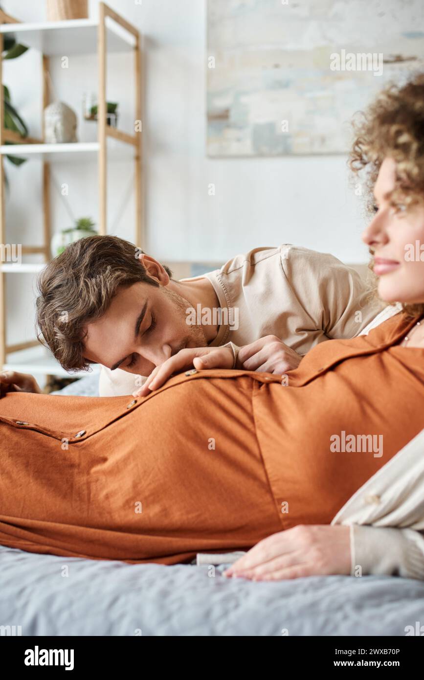 Mujer embarazada con el marido acostado en la cama, el hombre besando su vientre de la esposa y la mujer está sonriendo Foto de stock