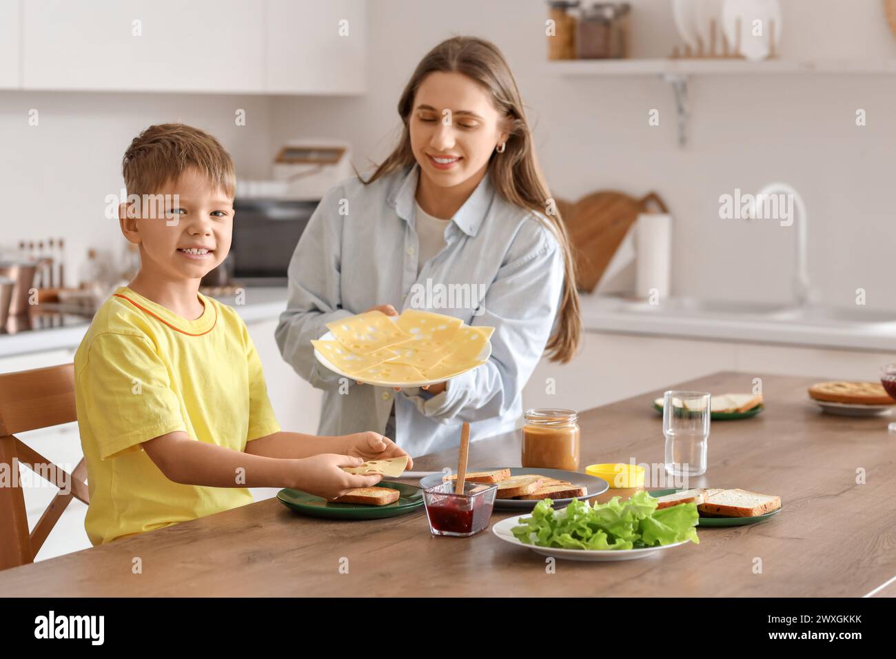 Lindo niño pequeño y su madre haciendo tostadas con queso en la mesa en la cocina Foto de stock