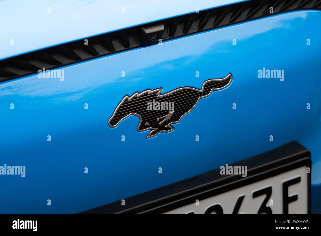 Emblema de Ford Mustang en la parte delantera de un coche eléctrico azul. Signo de logotipo con el caballo corriendo como parte de la marca automotriz. Foto de stock