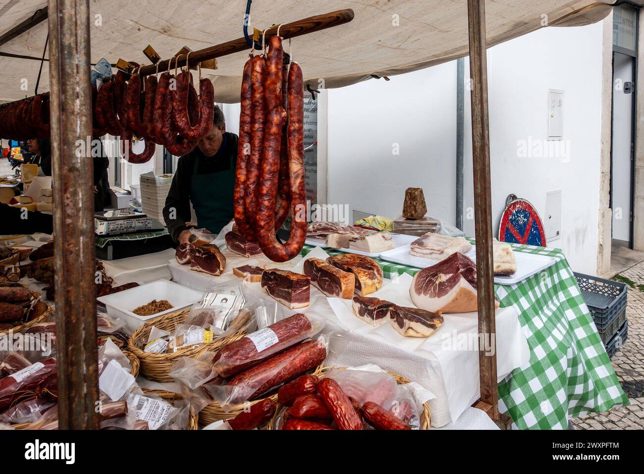 Un vendedor está detrás de un puesto adornado con una variedad de embutidos y embutidos, en Loule, Portugal. Foto de stock