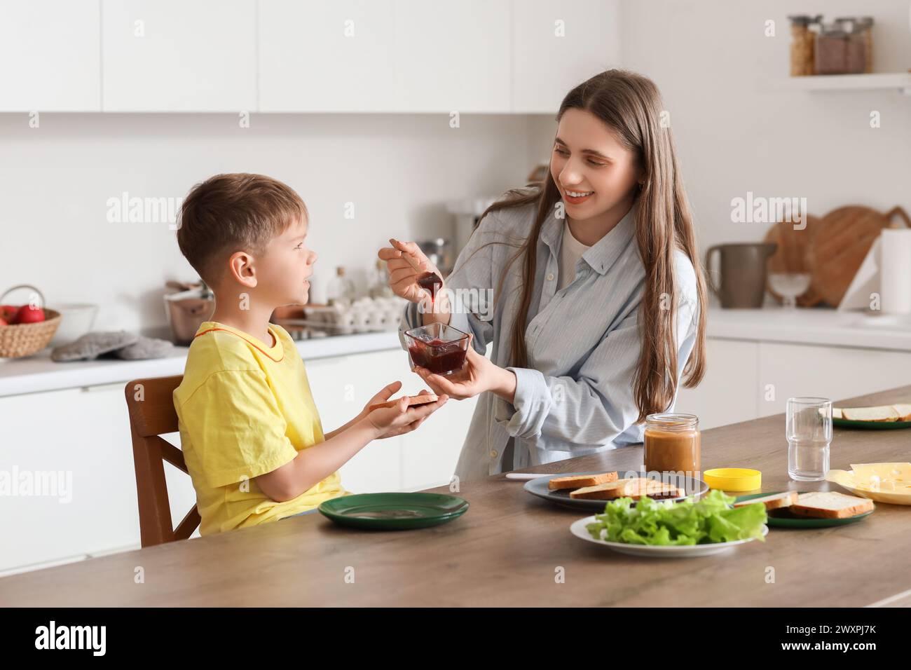 Niño pequeño con su madre haciendo tostadas con mermelada en la cocina Foto de stock