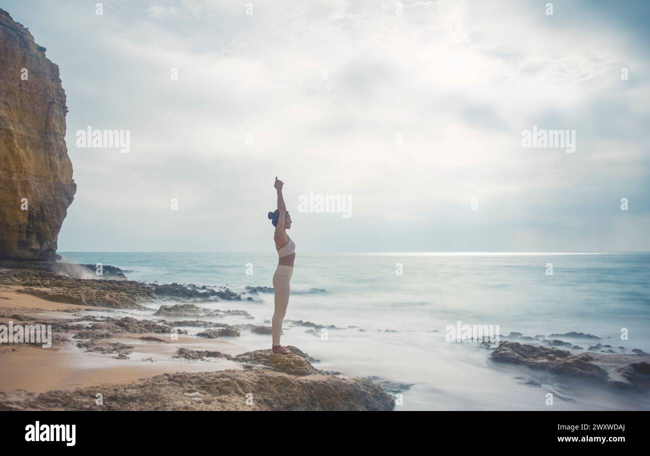 mujer de pie en las rocas junto al mar meditando y practicando yoga, los brazos por encima de la cabeza, larga exposición, mañana brumosa. Foto de stock