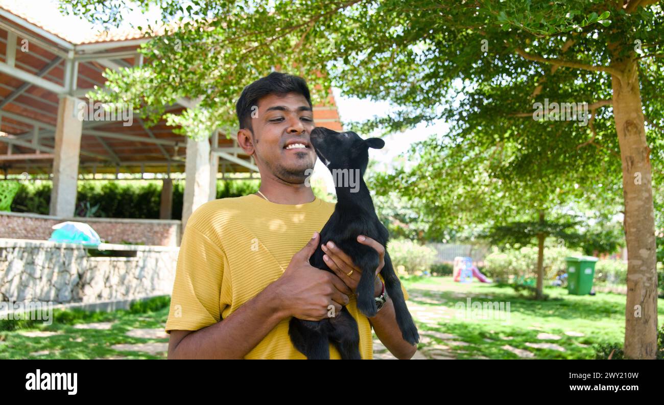 Un hombre indio gentil con una sonrisa cálida acuna a una pequeña cabra negra en sus brazos en un día soleado en un hermoso parque. Foto de stock