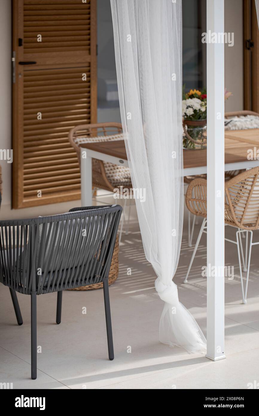 Un elegante patio al aire libre con una cortina blanca atada, sillas tejidas modernas y una mesa de madera, creando una atmósfera serena en un ambiente contemporáneo Foto de stock