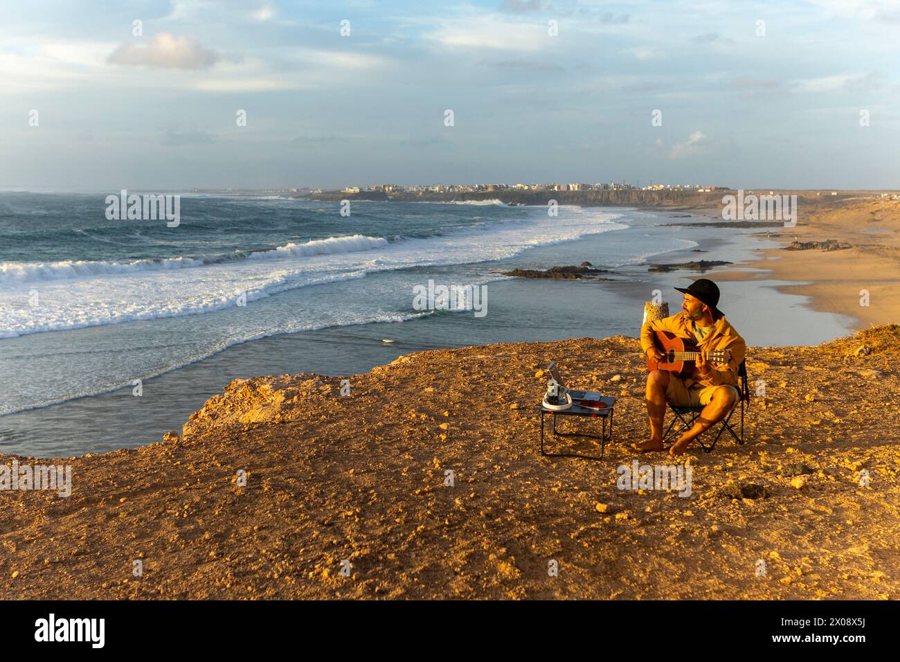 Un músico masculino, inspirado en las puestas de sol costeras, es visto componiendo en su guitarra junto al océano, viviendo un estilo de vida nómada fuera de su autocaravana. Foto de stock