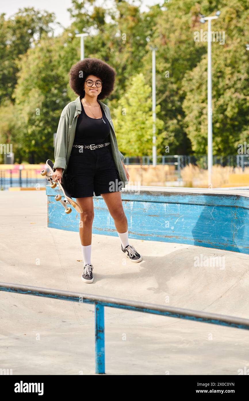 Joven mujer afroamericana con el pelo rizado sostiene una patineta, de pie junto a una piscina brillante. Foto de stock