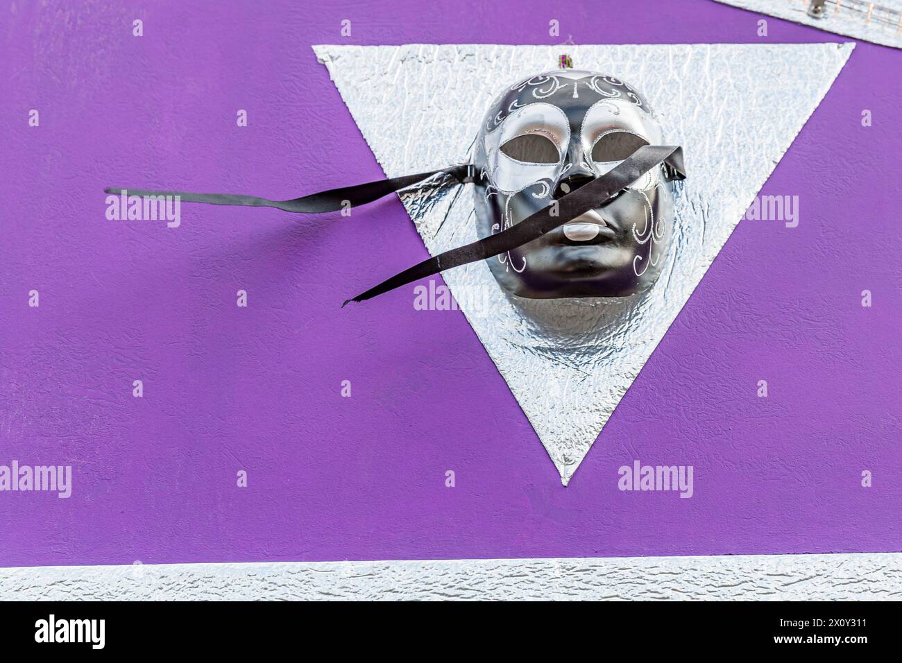 Máscara gris con detalles negros colgando en triángulo plateado en la pared exterior violeta, dos cintas negras sopladas por el viento, decoración en flotador de carnaval Foto de stock