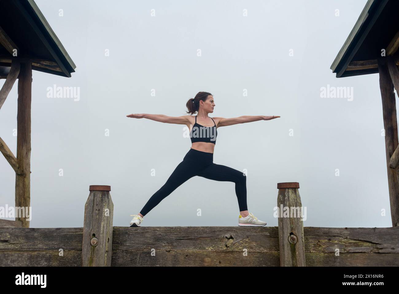 Mujer deportiva haciendo una pose de yoga guerrero, afuera por un edificio de madera Foto de stock