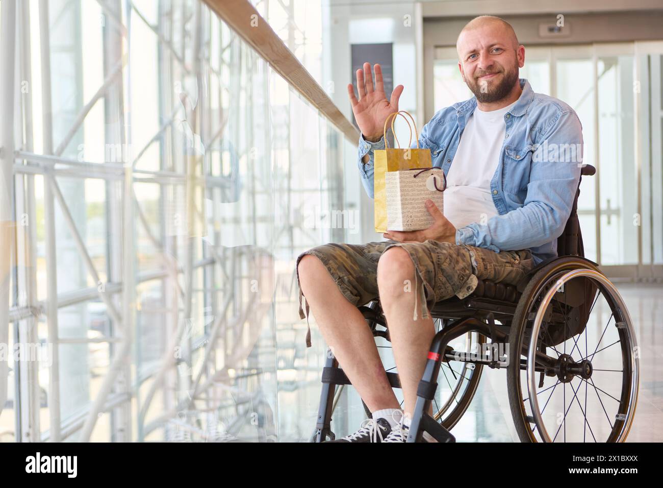 Persona alegre que usa una silla de ruedas ondea mientras sostiene un regalo, ejemplificando inclusividad y positividad en un entorno interior moderno. Foto de stock