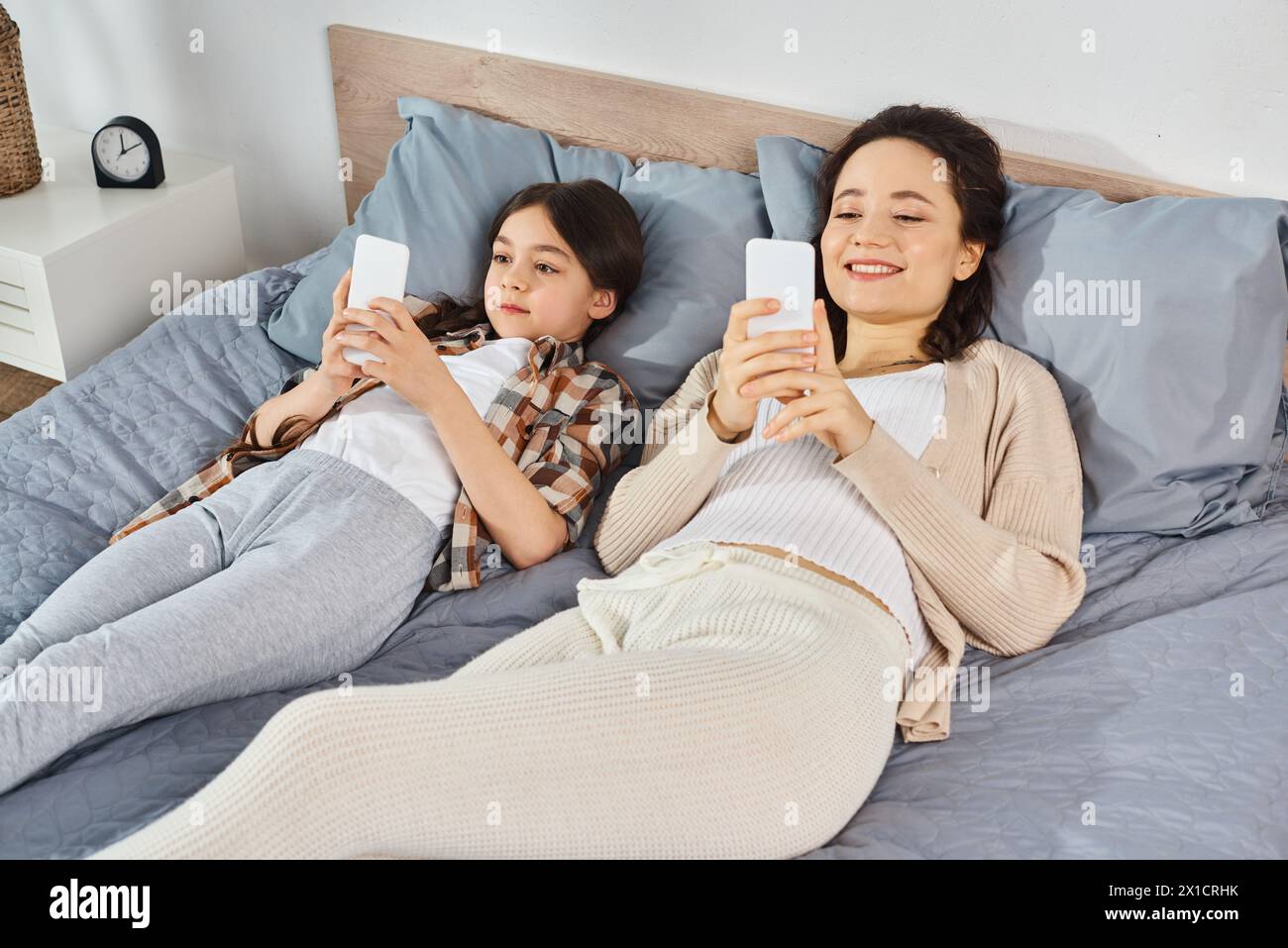 una madre e hija, acostados en una cama, sosteniendo teléfonos celulares y vinculándose con su tecnología. Foto de stock