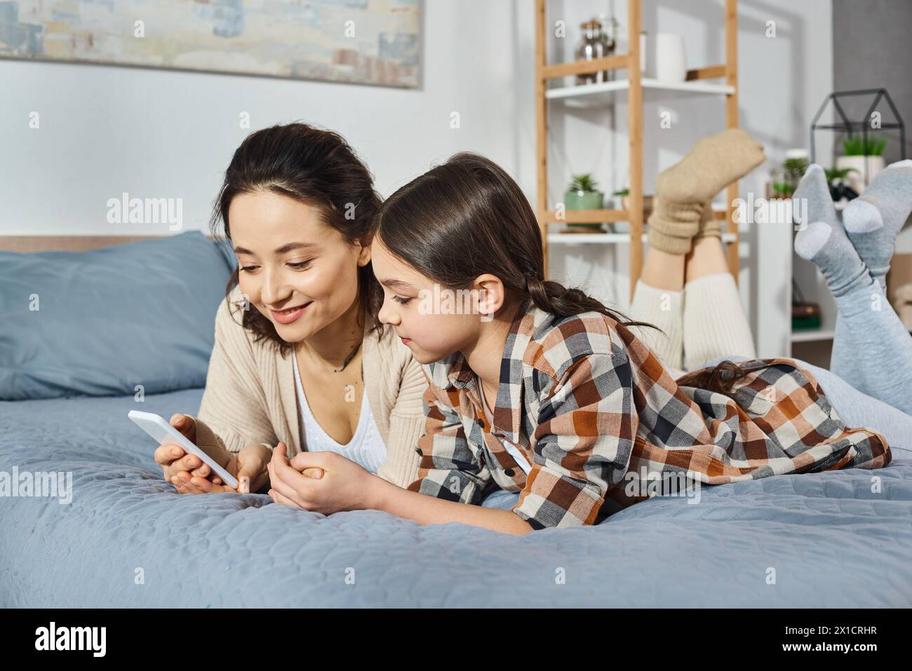 Una madre y una hija comparten un momento tierno mientras miran un teléfono celular mientras están acostados en una cama en casa. Foto de stock