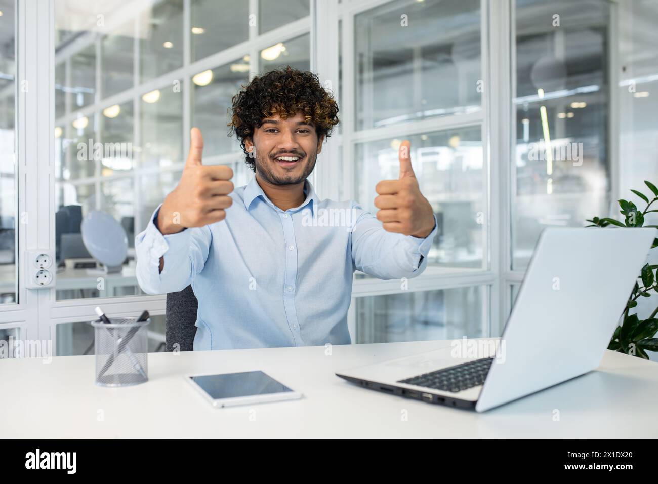 Un joven con una camisa azul claro sentado en un escritorio en un ambiente  de oficina brillante, dando con entusiasmo dos pulgares hacia arriba  mientras sonríe, retratando el éxito y la satisfacción